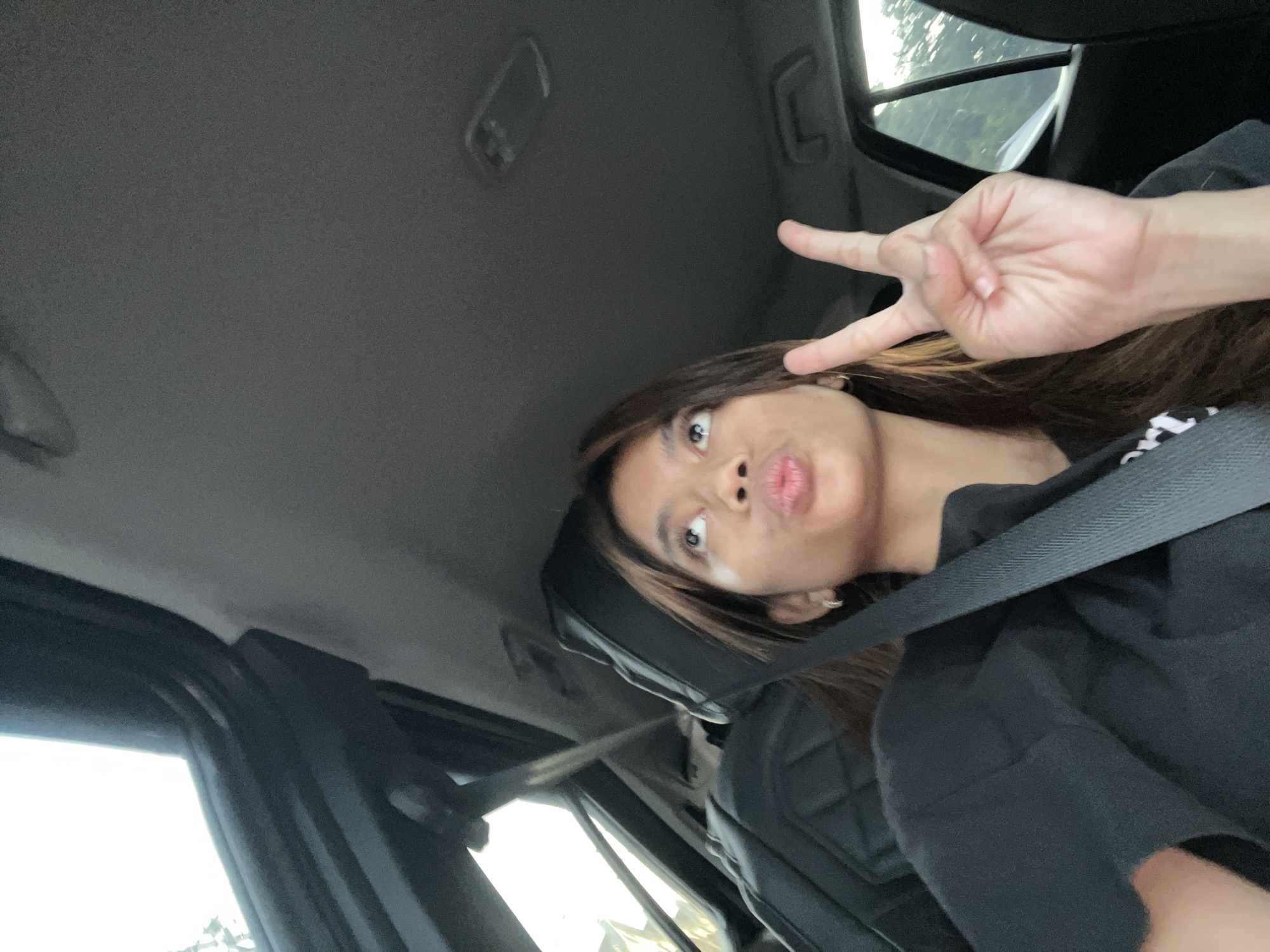 A young woman wearing a seatbelt is inside a car, possibly in Davao City, Philippines. She is making a peace sign with her right hand and pouting her lips. The car's interior is visible, including the ceiling, windows, and seats.