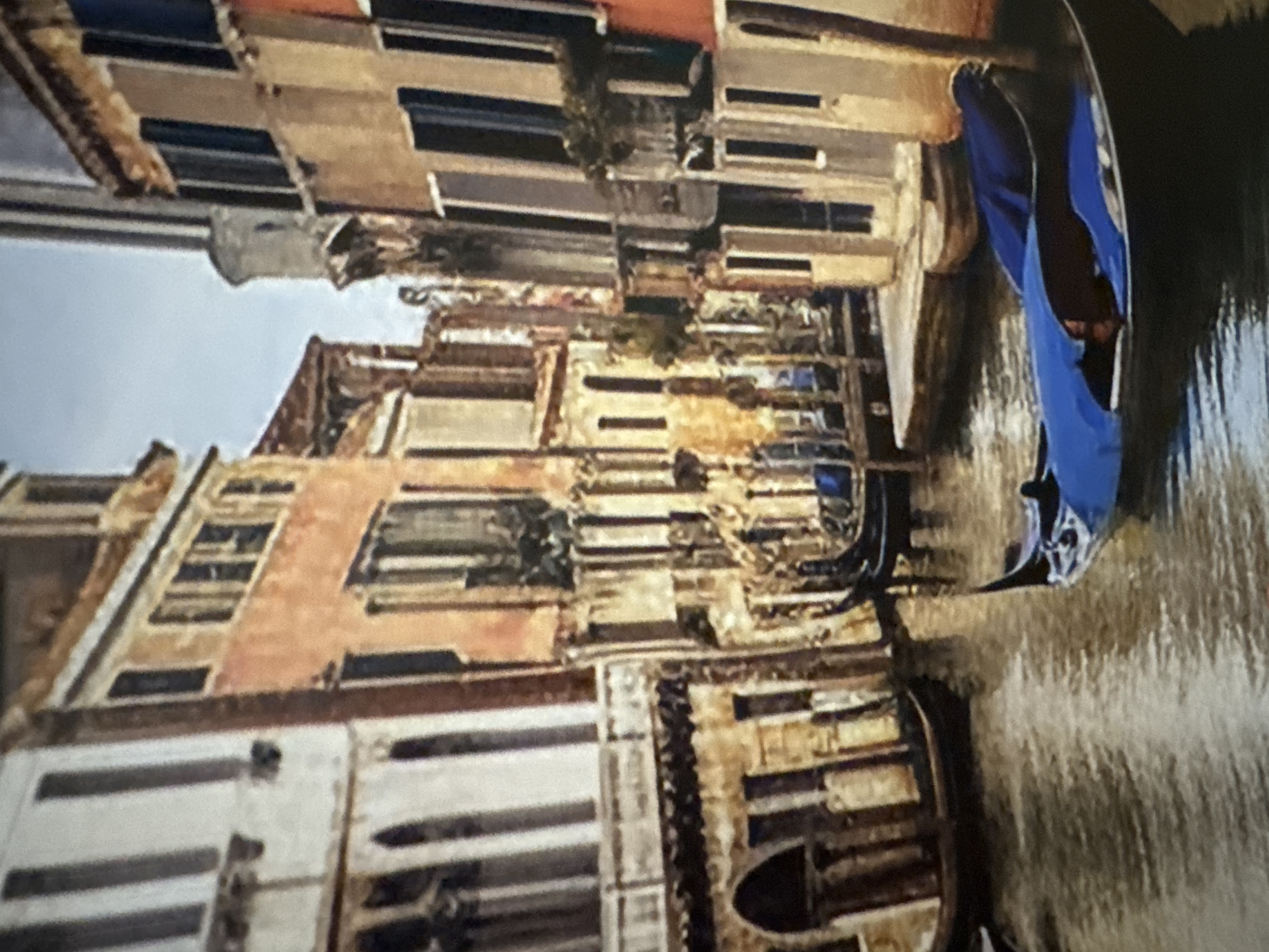 This is a painting of a canal in Venice, Italy. The canal is lined with tall, narrow buildings with many windows. In the foreground, a black gondola with a blue cover is floating on the water. In the background, another gondola is visible, also on the canal. The sky is overcast and a pale blue.