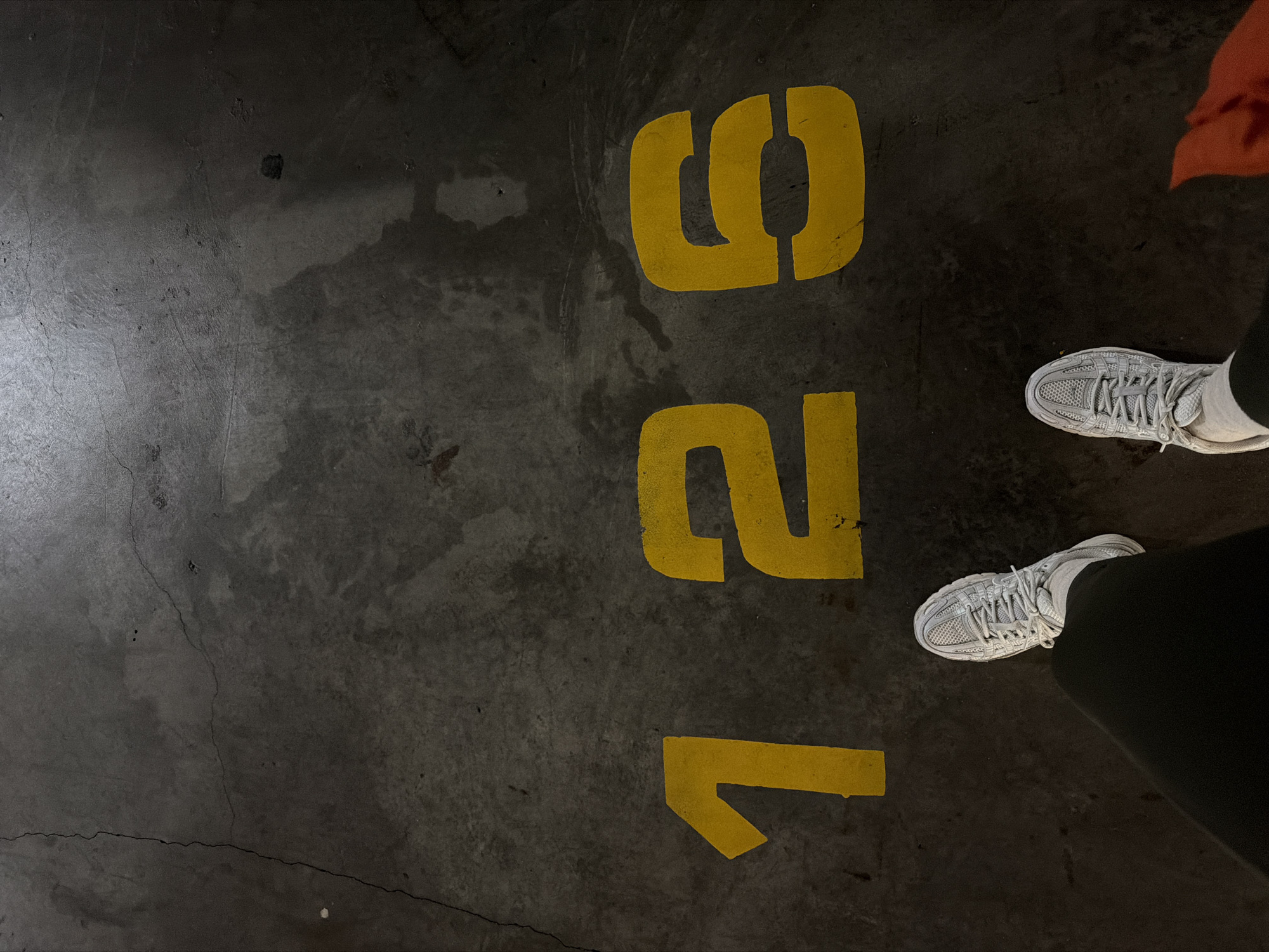 The image shows a section of a concrete floor, possibly in a parking garage or similar industrial setting. The floor is dark gray and worn, with various imperfections and cracks. Painted in yellow on the floor is the number "126," oriented vertically. The feet of a person wearing white athletic shoes and black pants are visible in the upper right corner. The presence of part of an orange garment suggests they are wearing a jacket or another layer of clothing of that color. The image does not contain sufficient information to confirm the location in Givatayim, Israel. The overall impression is of an unadorned, functional space.