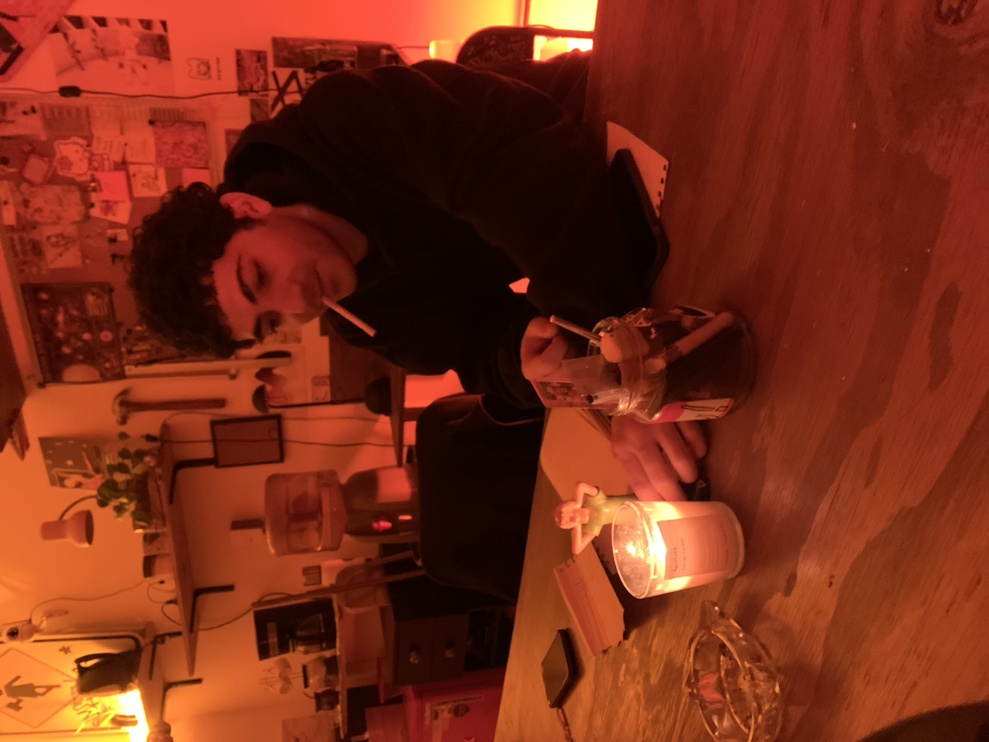 A person with dark curly hair, wearing a dark hoodie, is sitting at a wooden table in a room with warm, reddish lighting. The person has a cigarette in their mouth and is holding another small stick in their hand, near a glass jar containing similar sticks. On the table, there is a lit candle in a glass container, a small figurine, and a smartphone. In the background, there is a wall with various posters and items, a water dispenser, and other objects on shelves. The room is indoors and appears to be a living or recreational space. There are no indications of the location being in Buenos Aires, Argentina.