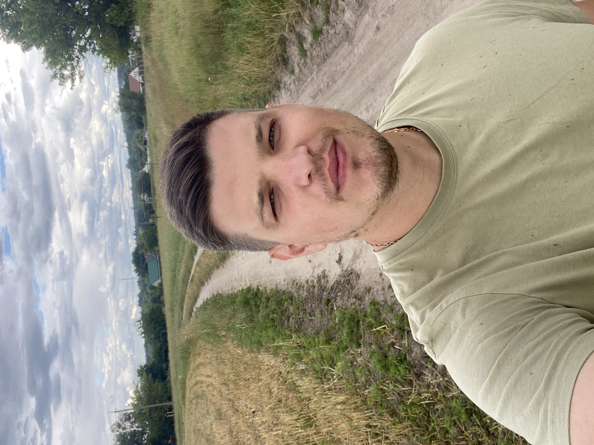 In Yablunivka, Ukraine, on a cloudy day, a man with short brown hair and a beard takes a selfie outdoors. He is wearing a light green t-shirt and a gold chain. The man is standing on a dirt road that winds through a field. To his left, the field is covered in cut straw, and to his right, the field is filled with tall green grass. In the background, scattered houses and trees are visible under the overcast sky.