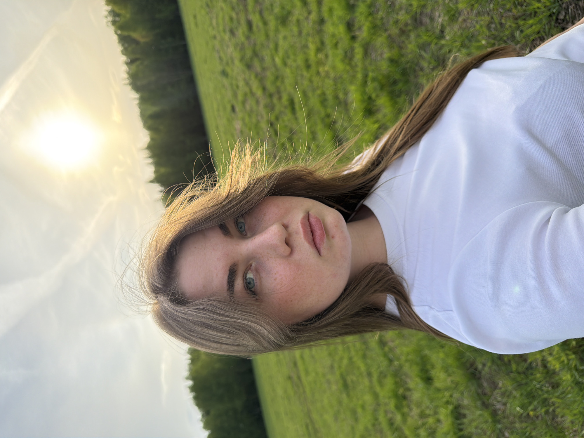 A young woman is lying on a grassy field. She has light brown hair, fair skin with freckles, and light-colored eyes. She is wearing a white shirt. The background shows a field with trees in the distance and a bright sun in the sky.
The image provides no information about the city of Zlodar', Russia.