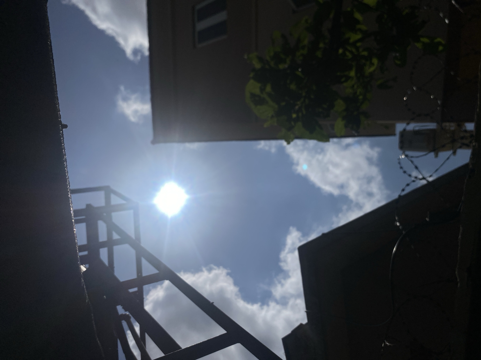 The image depicts a bright, sunny day in Barden Yana, Nigeria. The sun is prominently featured in the sky, creating lens flare and strong light. In the foreground, a dark, silhouetted structure, possibly a roof or overhang, extends into the frame from the top left. Below this, a metal structure, likely scaffolding or some form of construction element, is also silhouetted against the sky.
To the right of the frame, a portion of a building, possibly a multi-story structure, is visible. It appears to be constructed with a light-colored material. In front of this building, a dark green leafy plant or tree is partially visible, its foliage obscuring some of the building.
At the bottom of the image, a dark, silhouetted foreground element includes barbed wire, suggesting a boundary or security measure. The sky is blue with scattered white clouds. The overall scene is an outdoor urban environment bathed in bright sunlight.