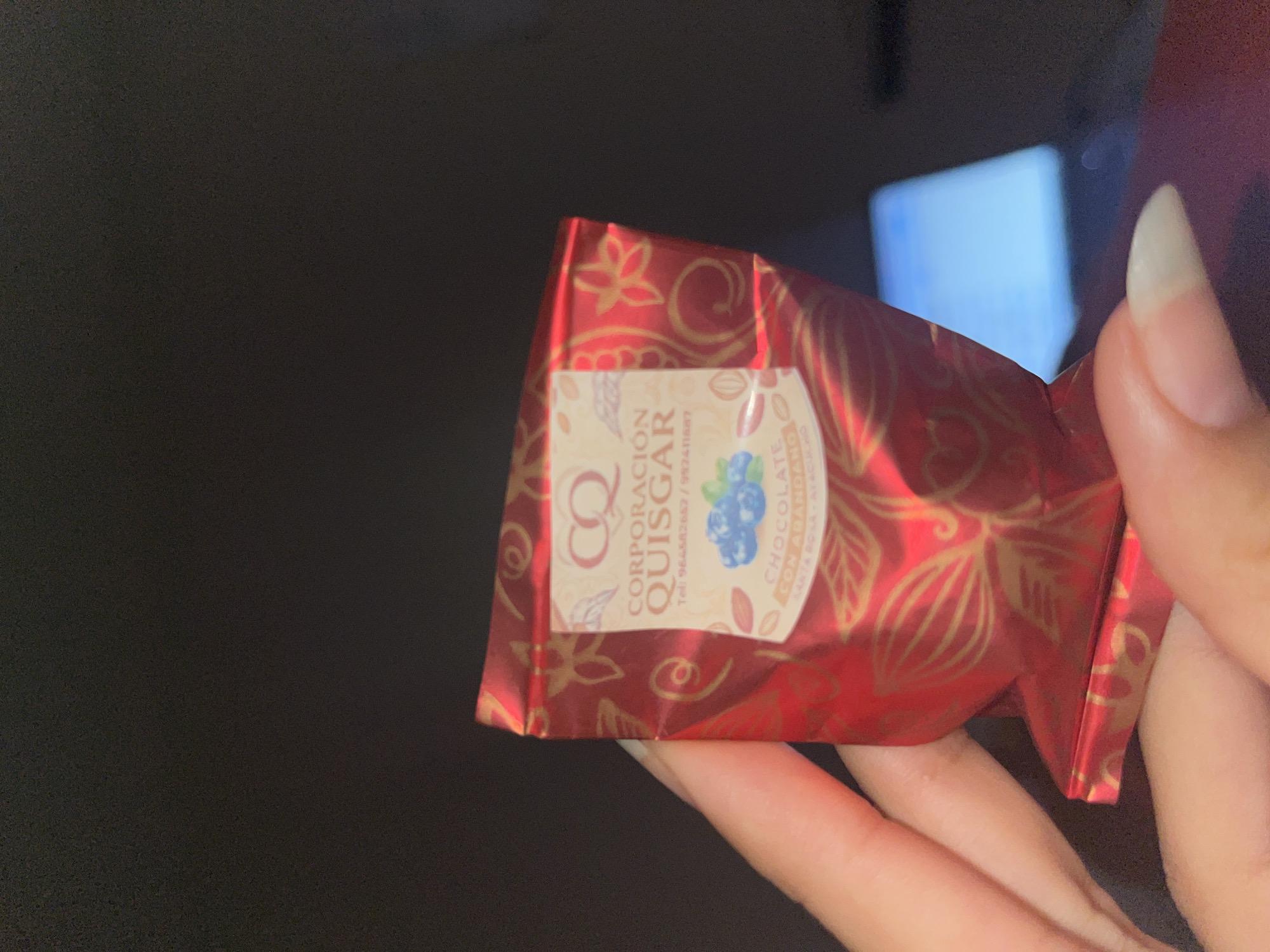 Here is a detailed description of the image:
A close-up shot features a hand holding a wrapped chocolate bar. The chocolate bar wrapper is red with gold designs and a label that reads "CORPORACION QUISGAR." The label also indicates that the chocolate is blueberry flavored. The background is dark and out of focus, showing a glimpse of a bright white screen in the distance. The focus is primarily on the chocolate bar and the hand holding it. There is no indication of the city San Luis, Peru in the provided image.