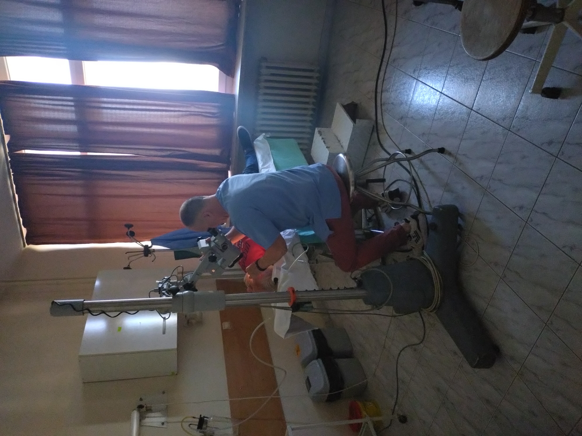 Location: Stara Zagora, Bulgaria.
A man in a blue shirt and red pants is using medical equipment in a room. The room has tiled floors, brown curtains, a radiator, and medical supplies. A patient is lying on a medical bed, being examined by the man.