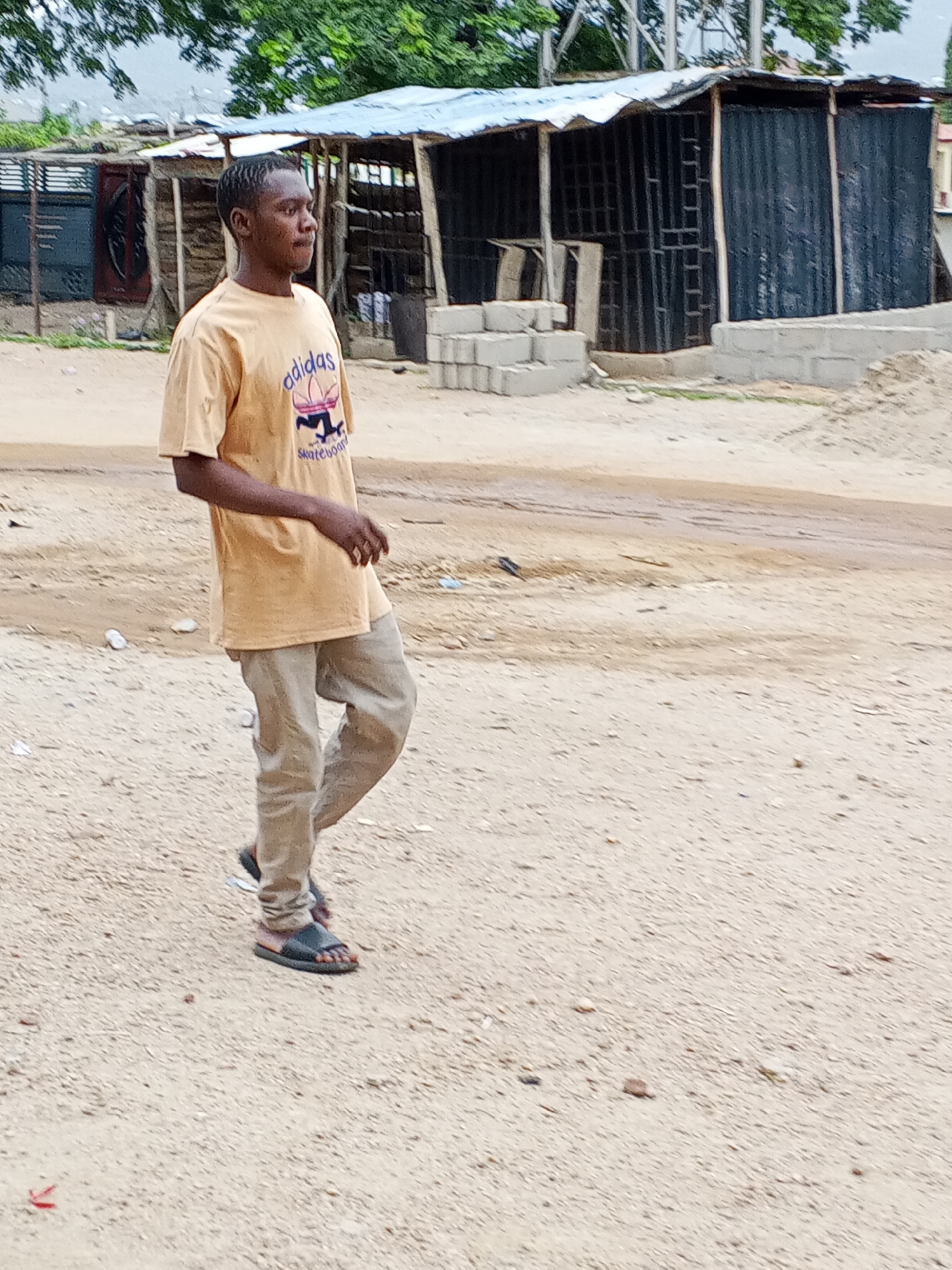 A young man is walking on a dirt road in Otokiti, Nigeria. He is wearing a tan t-shirt with an Adidas logo, tan pants, and sandals. The background consists of shacks constructed from corrugated metal and wood. There are concrete blocks and construction debris scattered around the area. A few trees and a mountain are visible in the distance.