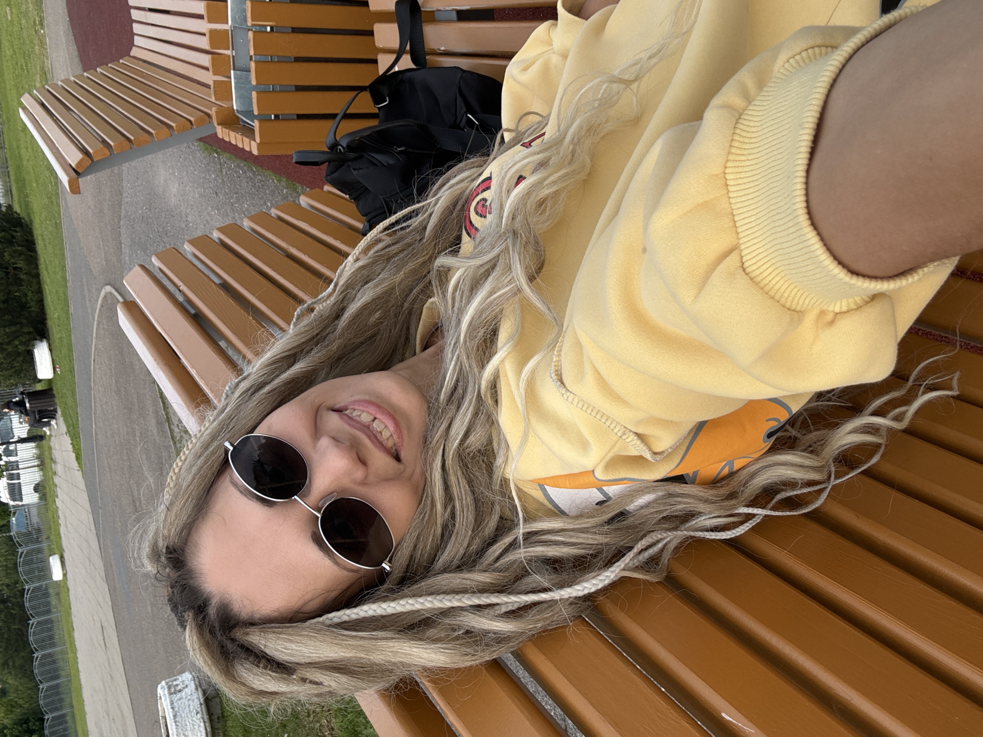 A young woman is captured in a casual outdoor setting, lying on her back on a wooden slatted bench and smiling broadly at the camera. She has long, wavy blonde hair, with several sections featuring lighter, braided extensions that resemble dreadlocks. Her eyes are covered by round, dark-lensed sunglasses with thin silver frames. She is dressed in a bright yellow hooded sweatshirt with ribbed cuffs, and an orange top with a circular graphic is partially visible underneath.
A black backpack is positioned on the bench behind her head. The benches are constructed from warm brown or orange-toned wooden slats, characteristic of public outdoor seating. The background reveals a paved path or road, a grassy area, and a metal fence, possibly indicating a park, recreational ground, or public square. Some vehicles can be vaguely seen in the far distance. The scene is illuminated by soft daylight, suggesting a pleasant time of day. The woman's relaxed posture and happy smile convey a sense of contentment. The setting is likely an outdoor public space in Шебунино, Russia. No legible text is visible in the image.