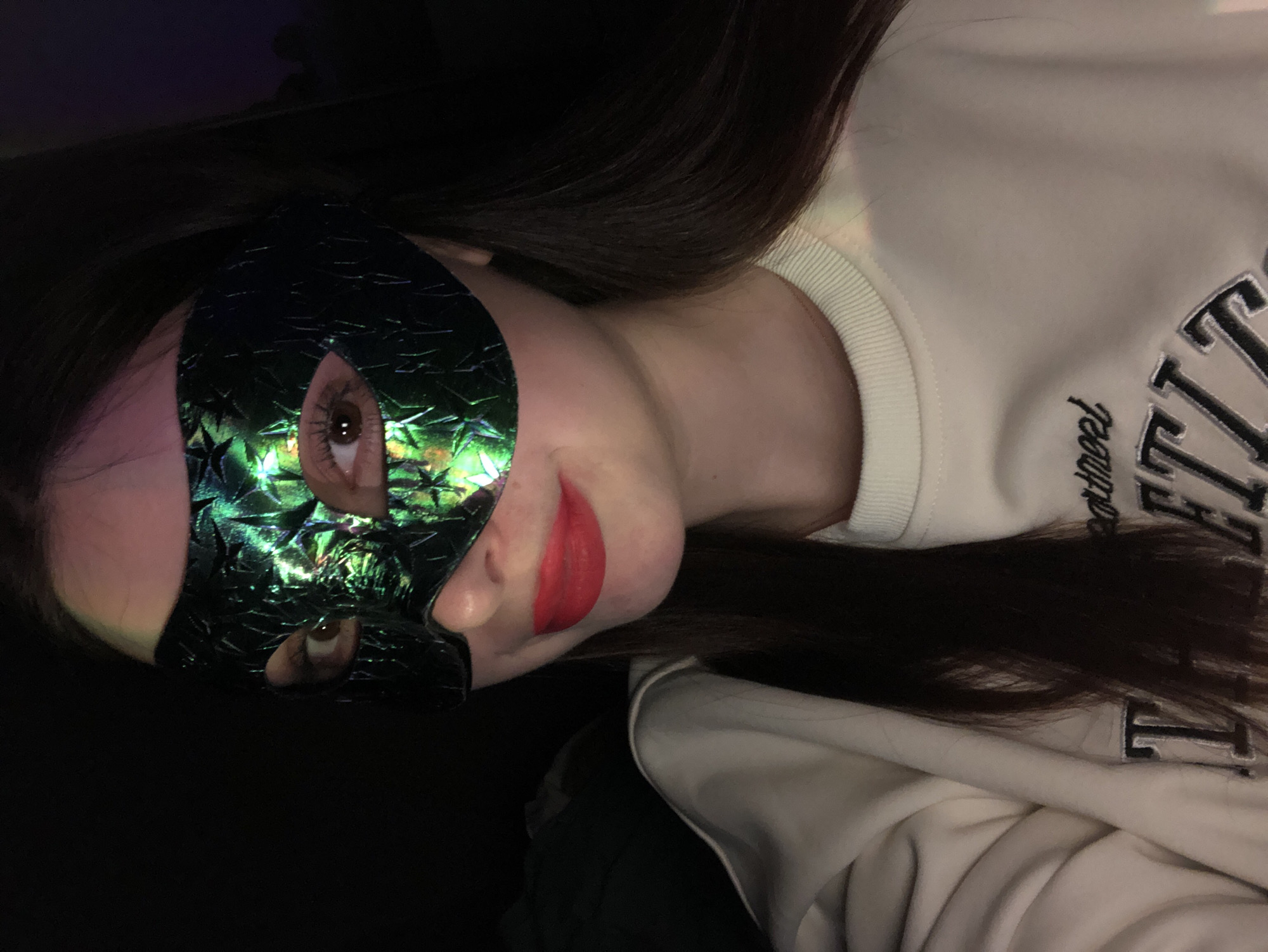 A woman is wearing a green and black iridescent masquerade mask that covers the left side of her face. Her hair is dark brown and long. She is wearing red lipstick. The rest of her face is visible. She is wearing a white sweater with a black letter design. The background is dark. There is no evidence to suggest this photo was taken in Noviy, Russia.