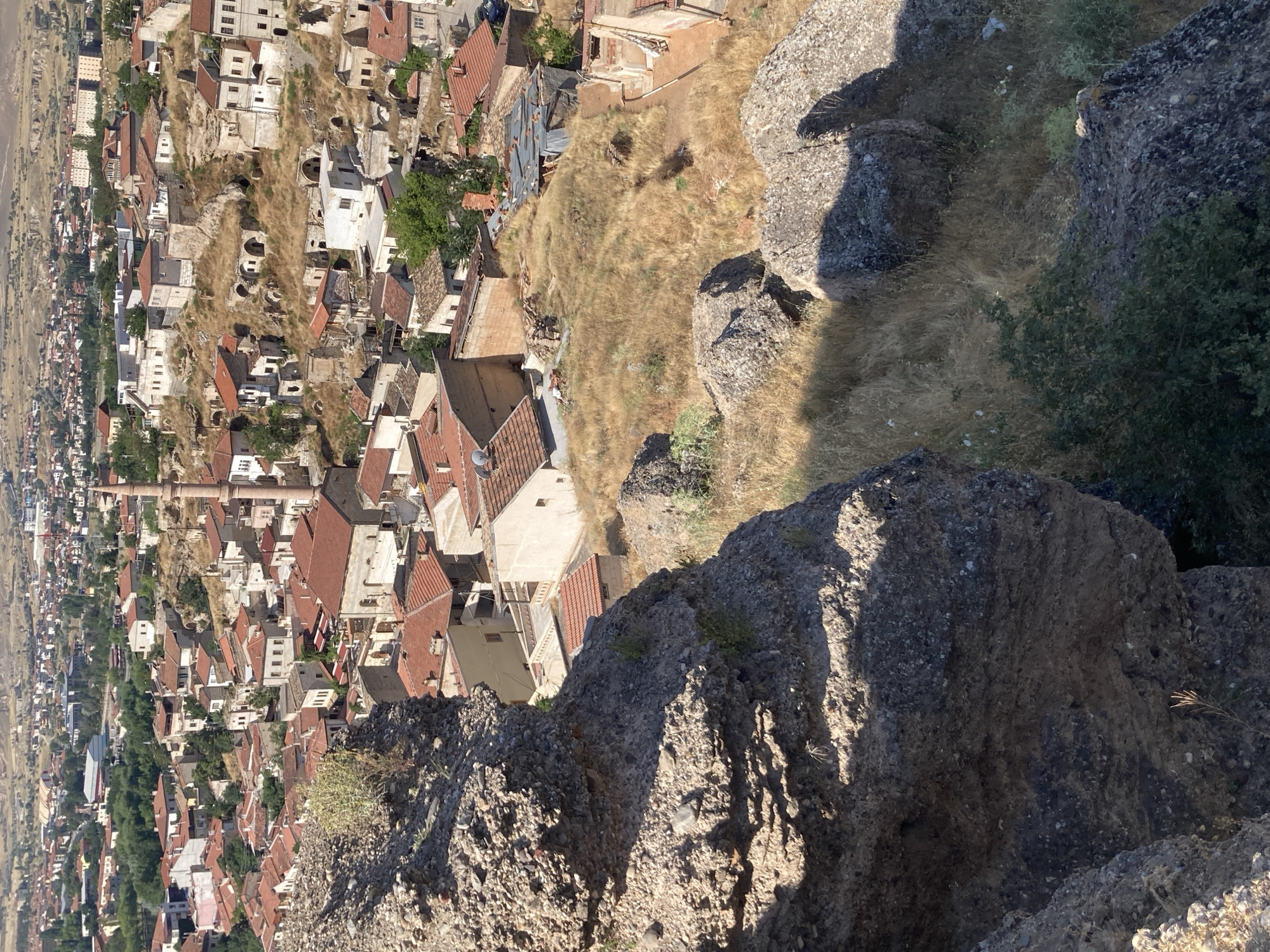 The image depicts a panoramic view of Avanos İlçe Merkezi, Turkey, from a higher vantage point. The foreground is dominated by rough, textured rock formations and dry, golden grasses. In the midground and background, a densely packed settlement unfolds, characterized by buildings with terracotta tile roofs. The architecture suggests a historical setting, with many structures appearing to be ancient or traditional. A prominent minaret rises from among the buildings, indicating the presence of mosques and an Islamic cultural context. The city is situated on a hilly terrain, with buildings cascading down the slopes. In the distance, more buildings and arid landscape are visible, stretching towards a hazy horizon under a bright, sunlit sky. The scene is devoid of any visible human activity.
