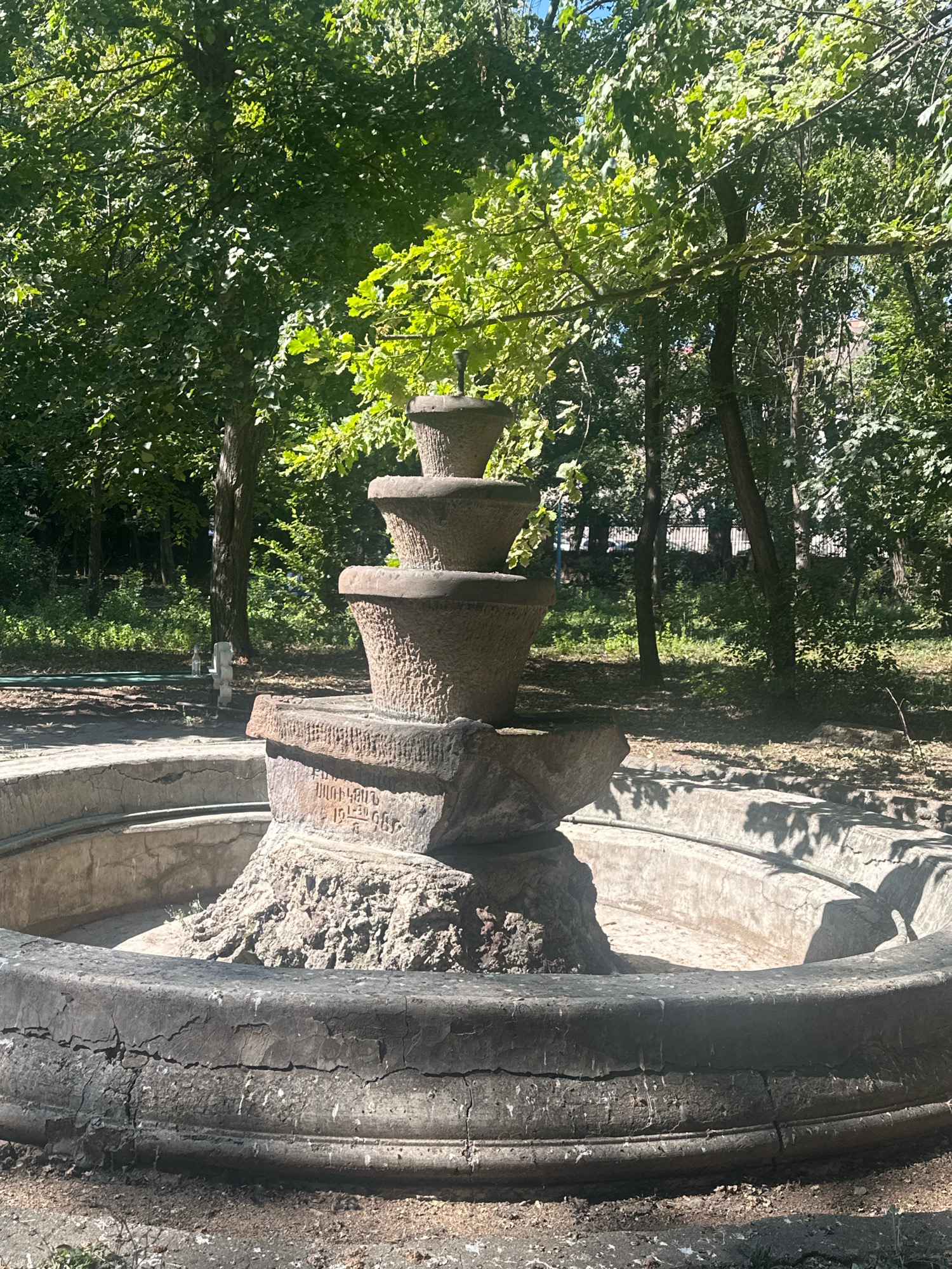 The image shows an old, multi-tiered stone fountain in a park-like setting. The fountain's central structure appears to be made of roughly carved stone and stacked basins, with a base of what looks like natural rock. The outer basin of the fountain is circular and made of concrete, showing signs of age with visible cracks and wear. Behind the fountain, lush green trees create a dense canopy, with sunlight filtering through the leaves, casting dappled shadows on the ground. The area around the fountain is covered in dirt and fallen leaves. In the background, a metal fence is visible, and beyond that, some buildings can be seen through the trees. The scene is set in the city of Gyumri, Armenia, likely in a public park or garden. There are no people or animals visible in the image. The overall impression is one of a somewhat neglected but charming historical site.