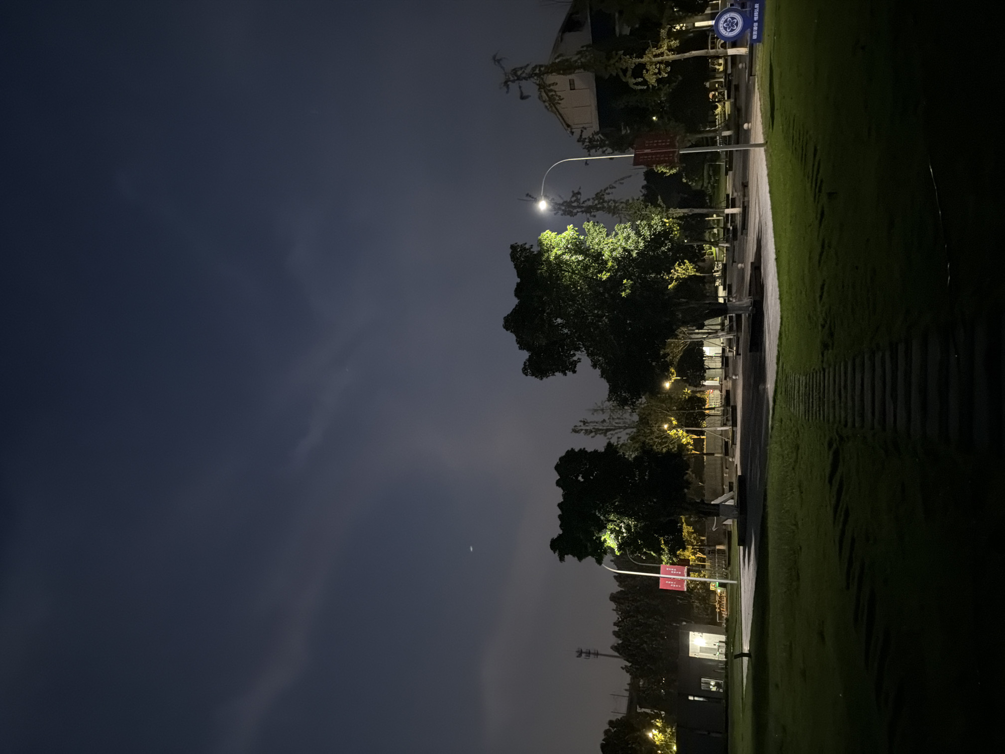 This image depicts a night scene in the city of Jiangjin, China. A pathway made of stone slabs leads through a grassy area towards a paved walkway. Two trees are illuminated by spotlights, one on the left and another on the right. Beyond the trees, streetlights cast a glow on the surroundings. In the background, several buildings are visible, some with illuminated windows. A streetlamp stands prominently in the middle ground. The sky is dark with a few wispy clouds and a faint celestial object, possibly a star or a distant light, is visible. There are no people present in the scene. The overall atmosphere is tranquil and nocturnal.
