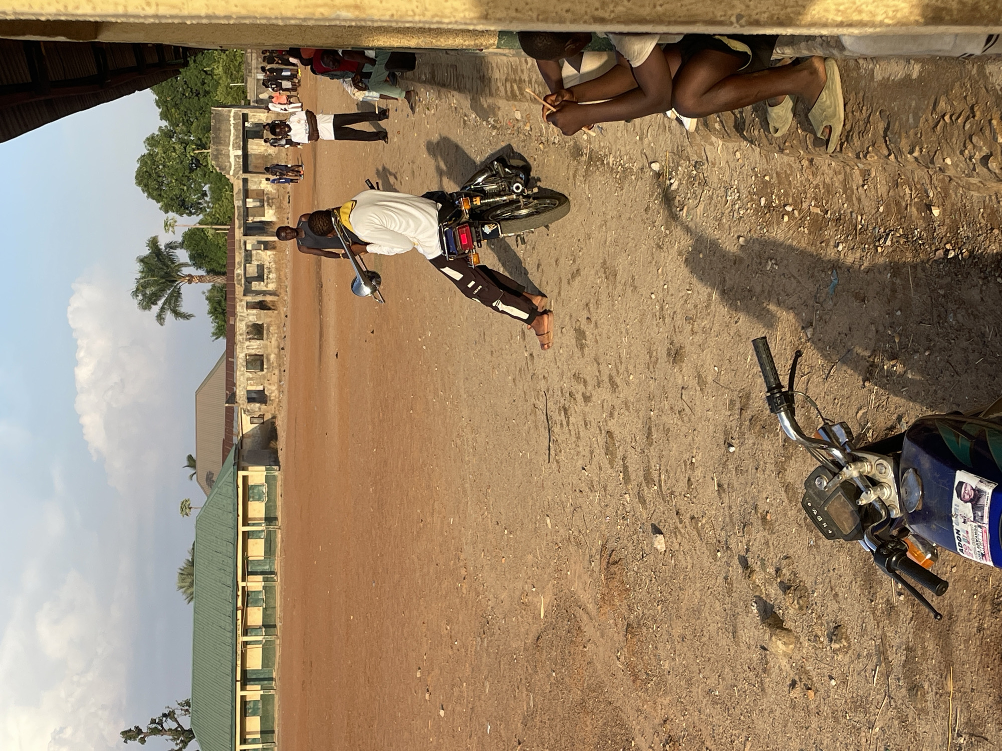 The image depicts an outdoor scene in Akwanga, Nigeria. The ground is dirt-covered. Several people are visible, including one individual in a white shirt and black pants holding a trombone while standing next to a motorcycle. Other people are standing in the background near a building with multiple windows. Another motorcycle is in the foreground. Two people are sitting. A palm tree is visible in the background against a partly cloudy sky.