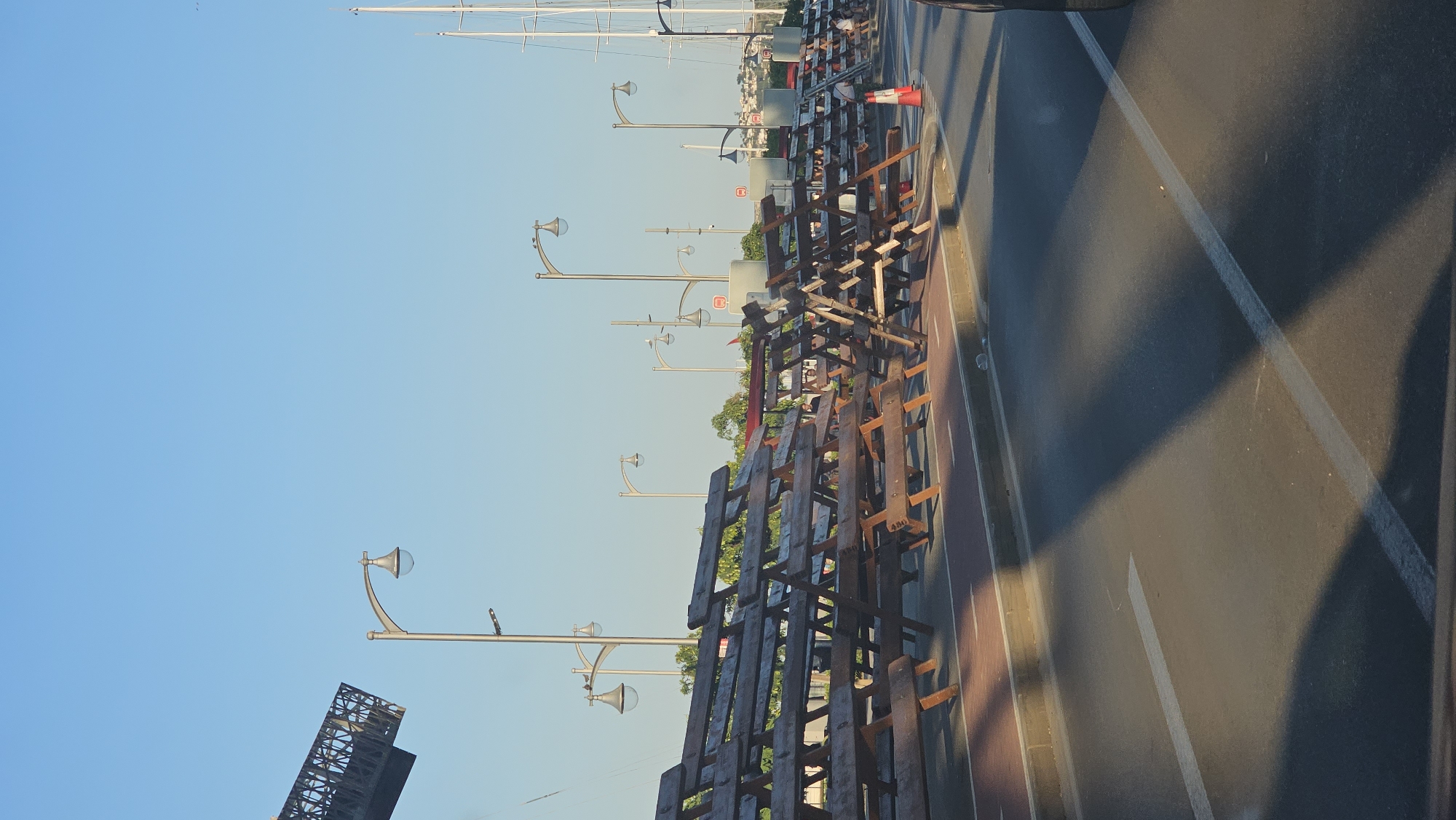 The image shows a section of the promenade in Dénia, Spain, featuring rows of wooden bleachers set up along the sidewalk adjacent to a road. The bleachers are arranged in a staggered pattern, creating a tiered seating area. In the background, several streetlights with modern designs illuminate the path. A clear blue sky dominates the upper portion of the image. To the right, the masts of sailboats are visible, suggesting the proximity of a marina. A traffic cone and what appears to be construction ladder are also present, indicating some ongoing work or event preparation. A person is partially visible near the ladder, and a few more individuals are scattered further down the promenade. The road in the foreground has white lane markings and the shadow of the bleachers casts a prominent pattern on the asphalt.