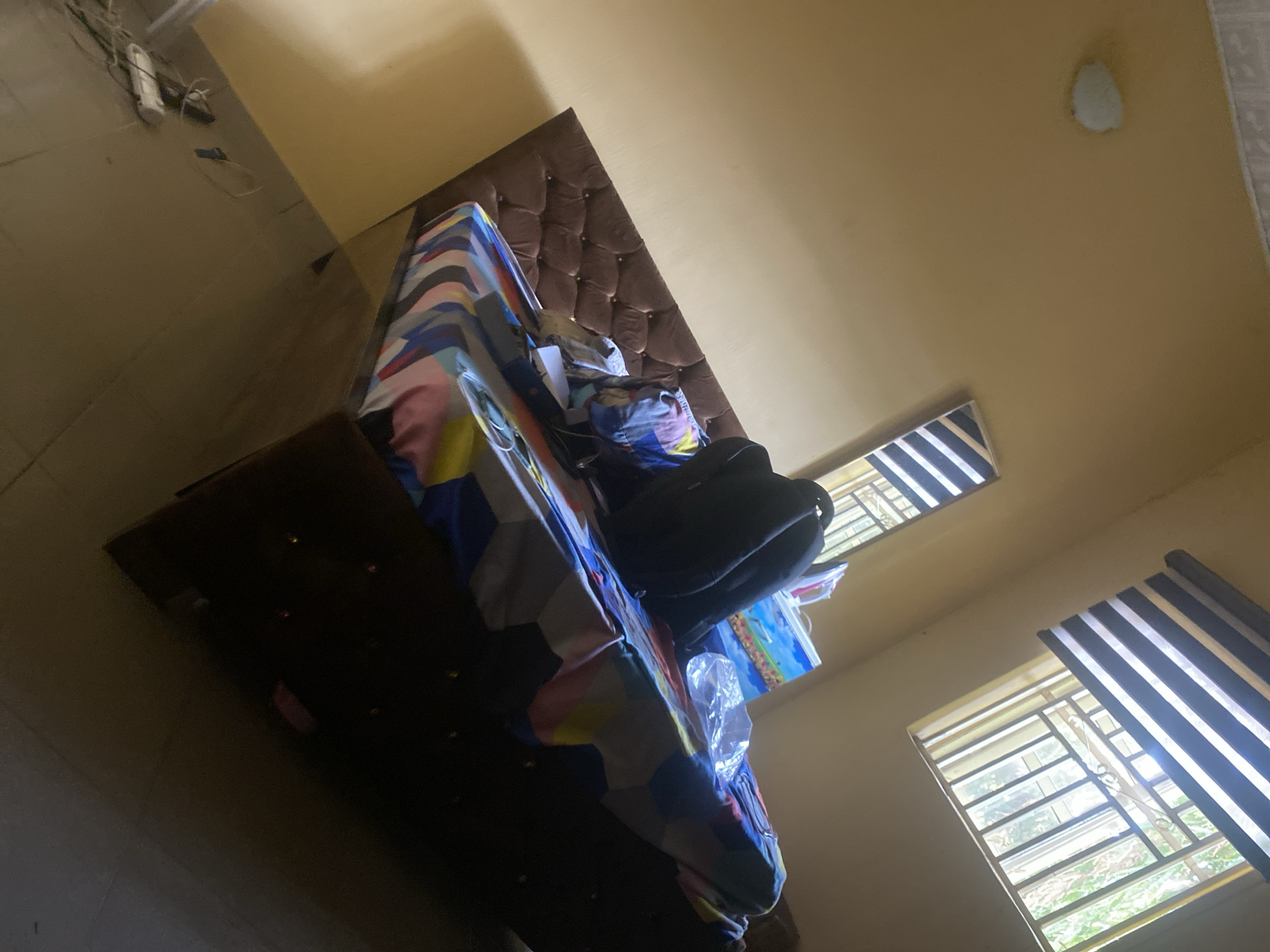 The scene depicts a bedroom interior in Hanwa, Nigeria. The primary feature is a bed with a tufted headboard, covered with a geometrically patterned bedspread. A black backpack is placed on the bed, along with other items. The room is lit by natural light streaming through two windows, one with striped blinds. The walls are painted a light yellow color. Electrical wiring is visible in the corner of the room.