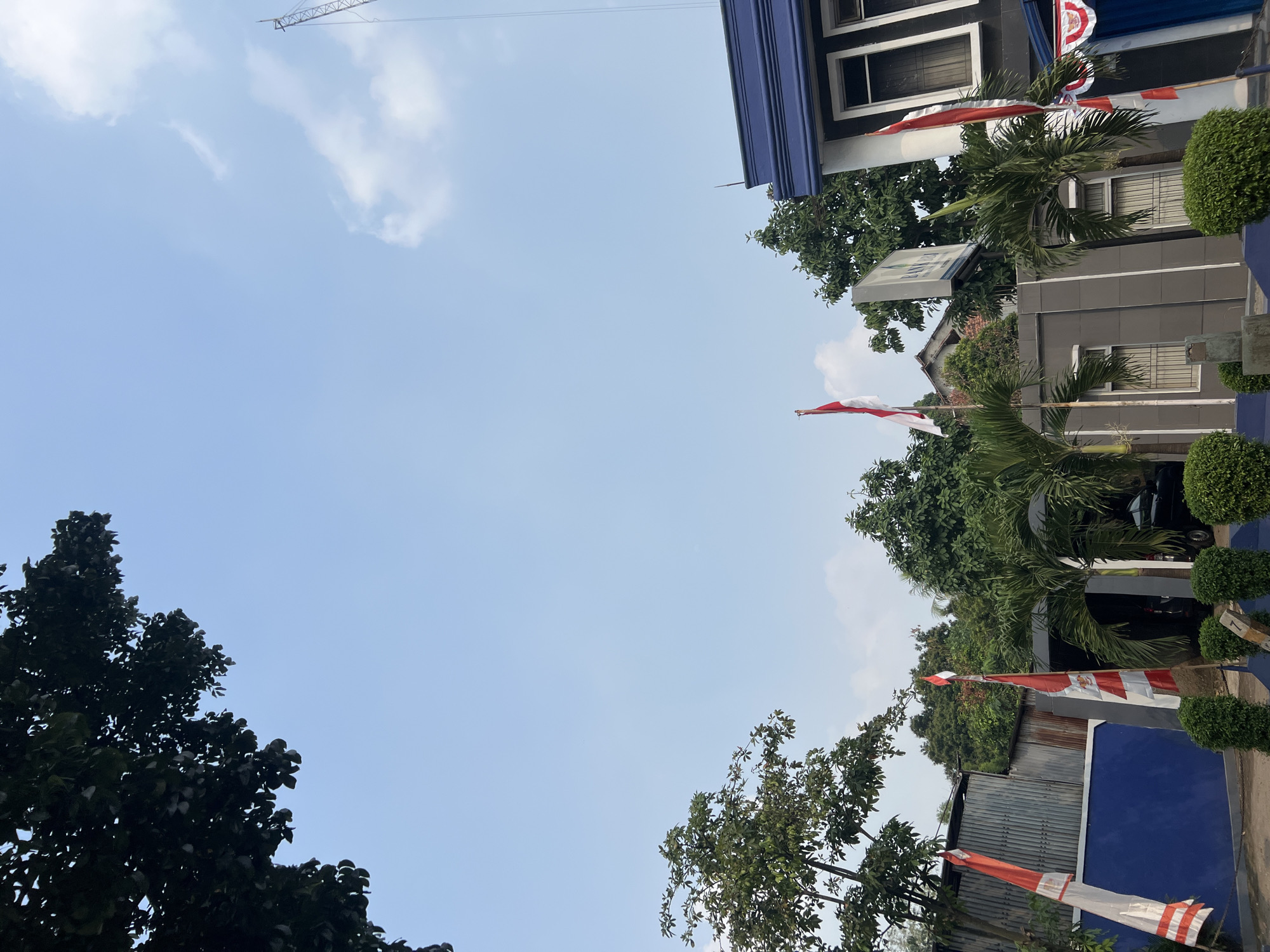 The image depicts an outdoor scene under a clear blue sky with scattered white clouds. In the foreground, there is a modern building with dark blue trim and white window frames. Several Indonesian flags, red and white, are prominently displayed on poles in front of the building. Lush green trees and palm trees surround the building, and a corrugated metal fence is visible on the left side. The scene appears to be in an urban or semi-urban setting. Based on visual cues, such as the architecture and vegetation, and common imagery associated with the region, the location is likely Citeureup, Indonesia. No people are visible in the image.