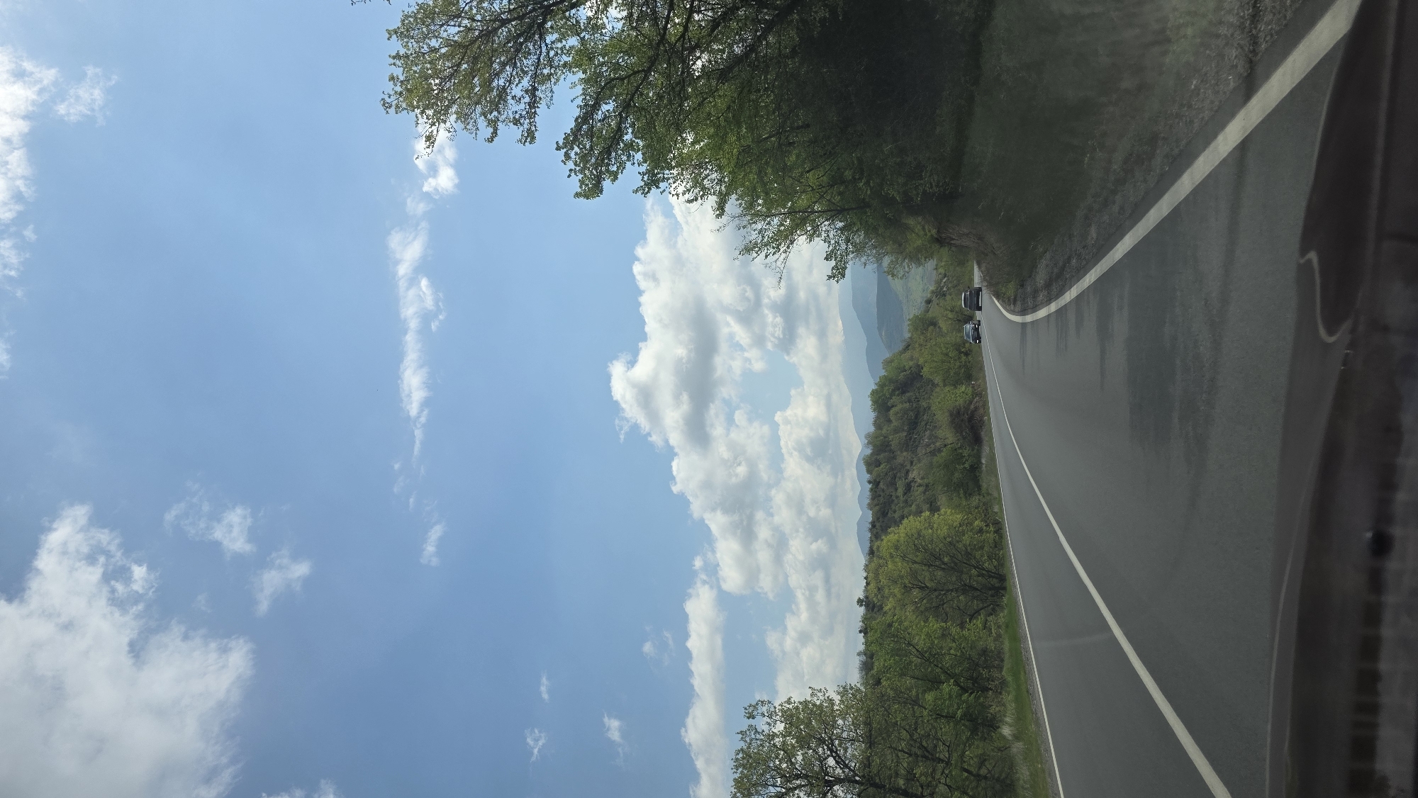 A road winds through a landscape with lush greenery and a bright, partly cloudy sky. The road is wet, suggesting recent rainfall. Two vehicles are visible on the road in the distance. The location is indicated as Vrania, Bulgaria, but there are no specific city details visible in this image.
