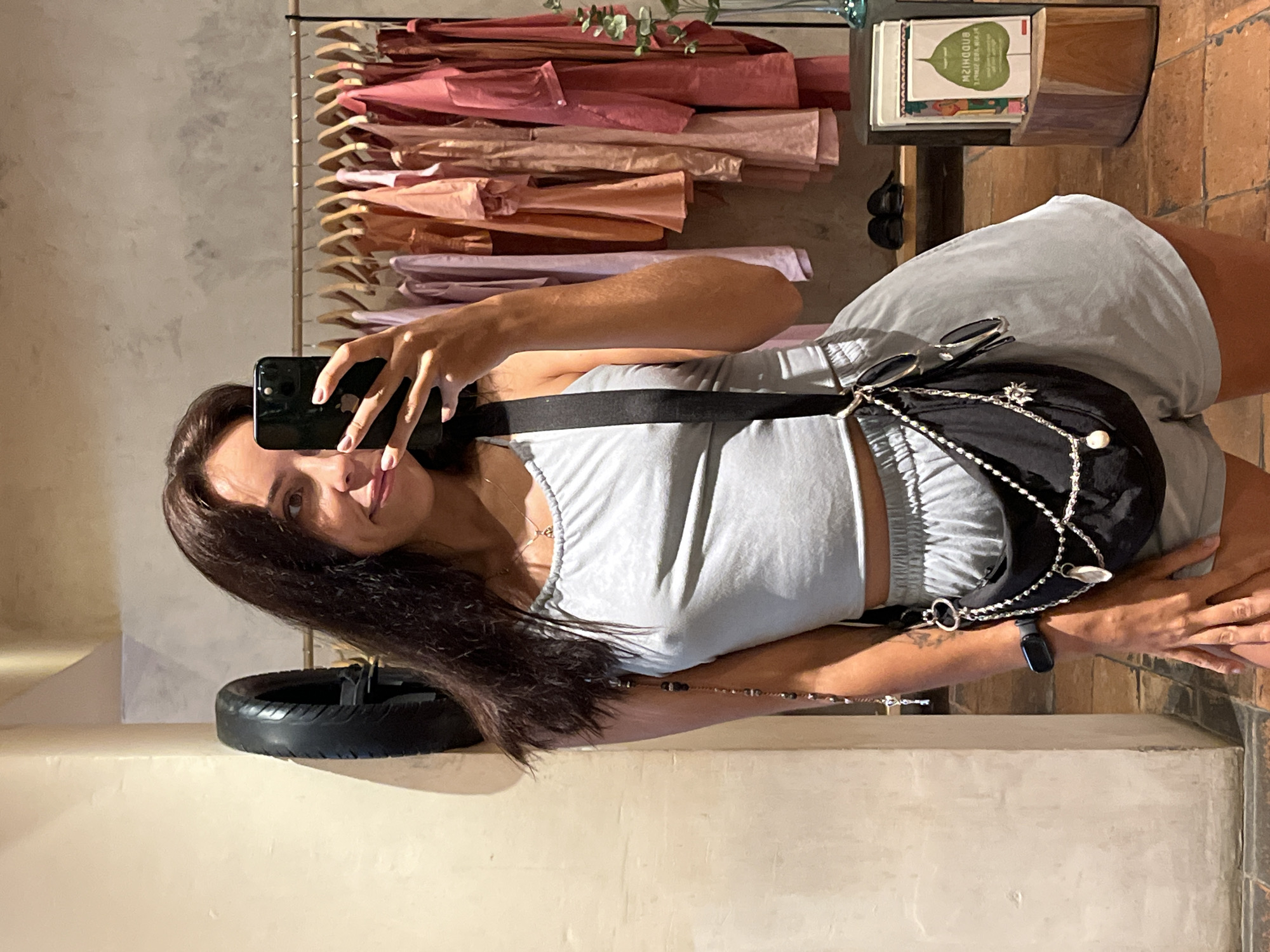 A woman is captured taking a mirror selfie indoors. The image shows her from the waist up, smiling faintly, with dark brown hair flowing over her shoulder. She is wearing a light grey two-piece outfit consisting of a cropped tank top and elastic-waist shorts. A black shoulder bag with silver chain details and white pearl accents is worn across her body, with a pair of sunglasses resting on top of it. A black fitness tracker is visible on her right wrist. She holds a black smartphone to capture her reflection.
To the viewer's left of the woman, a clothing rack is visible, displaying numerous folded or draped garments in various shades of pink, rose, and beige, suggesting a retail or storage area. To her right, a small wooden table holds a stack of books, one of which clearly shows the title "Buddhism" on its spine. Below the table, there's a dark, circular object that resembles a tire or a decorative tire-shaped stand. The floor beneath the objects is composed of terracotta-like tiles, while the walls are light-colored, some appearing to have a textured or concrete finish. Small green plant foliage can be seen near the top right, adding a touch of nature to the indoor setting. The lighting appears to be natural, suggesting daytime. The overall atmosphere is casual and relaxed.
