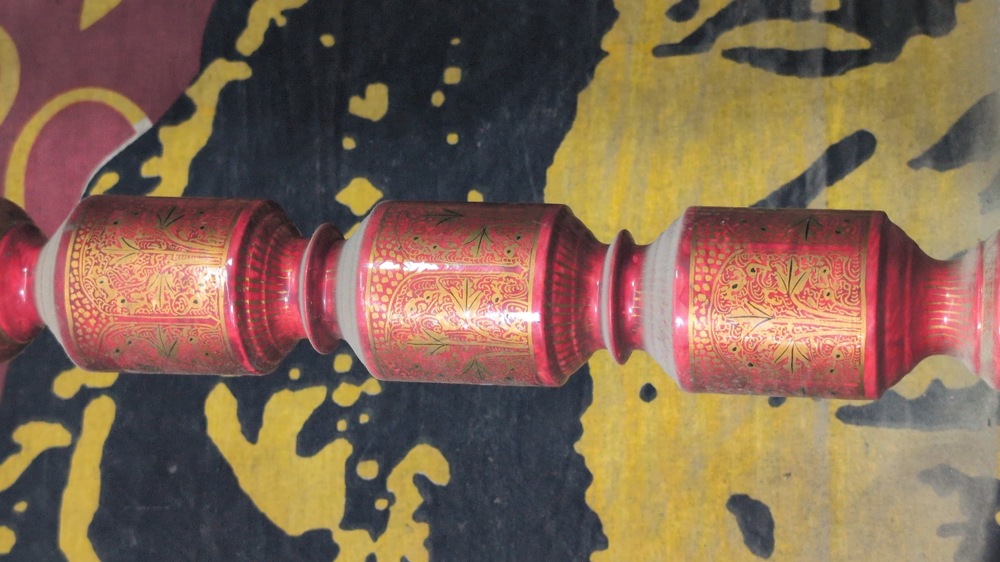 A vertical, close-up, eye-level shot shows a red, ornate decorative object with gold detailing, positioned in front of a textured background of black and yellow. The object is comprised of three stacked cylindrical segments, each adorned with intricate patterns resembling trees and geometric designs in a shimmering gold finish. The red surface of the object appears dusty. The background, a tapestry or fabric, features abstract, uneven shapes in yellow on a black base, with a maroon border visible at the top. The lighting is moderate, casting soft shadows and highlighting the textures of both the object and the background. There are no people or visible actions in the frame.