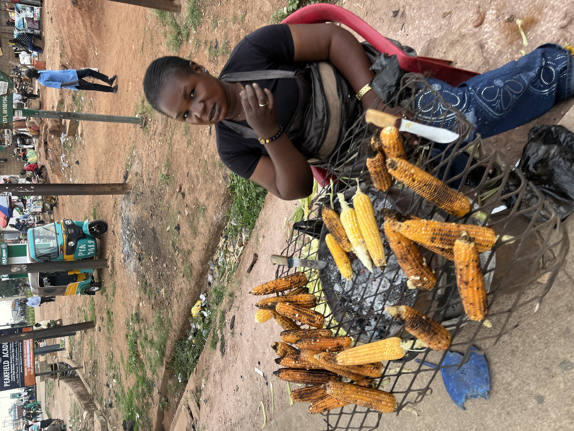 The image shows a woman selling roasted corn on the street in Gura Top, Nigeria. She is sitting on a red plastic chair next to a metal grill filled with roasting corn. Some of the corn is still yellow, while other pieces are roasted to a dark brown color. A white knife rests on the grill. More cooked corn is laid out on the ground next to the grill. The woman is wearing a black shirt, blue jeans decorated with white embroidery, and jewelry, and she is looking at the camera.
In the background, there is a street scene with people, vehicles (including a green and yellow tuk-tuk with the license plate "PTA 7419"), and buildings. One sign that's visible in the background advertises "PEAKFIELD ACADI" (likely an abbreviation of "academy"). There is trash in the gutter and general signs of a busy roadside environment.