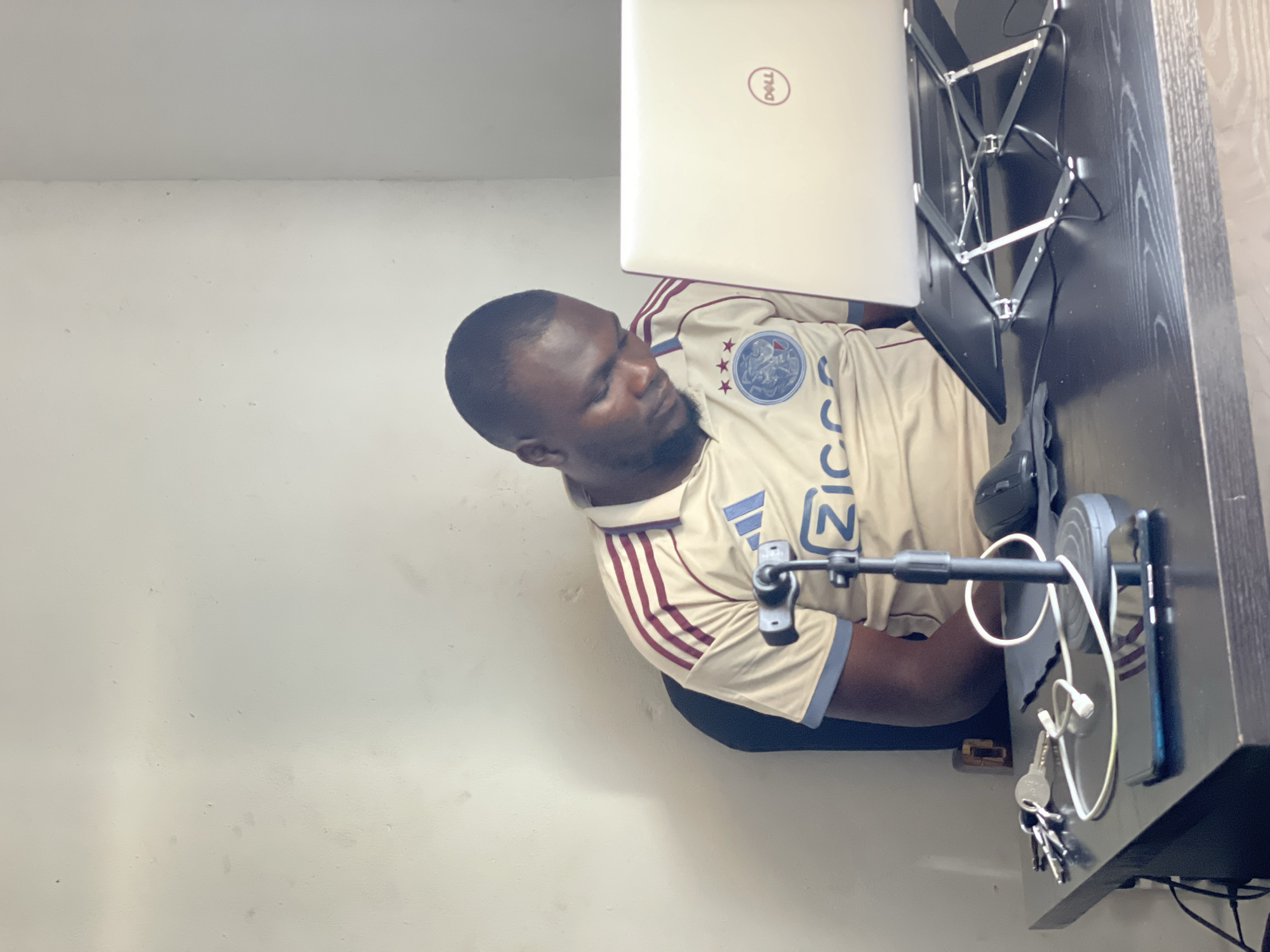 A man wearing a light brown t-shirt with red and blue stripes and an Ajax logo is sitting at a black desk in front of a laptop. The man appears to be looking down at the laptop, with his eyes closed. On the desk, there are several objects, including a microphone on a stand, a computer mouse, a smartphone, and a set of keys. The background of the image is a plain white wall. The scene is set in a room, likely a home office, in the city of Jos, Nigeria.