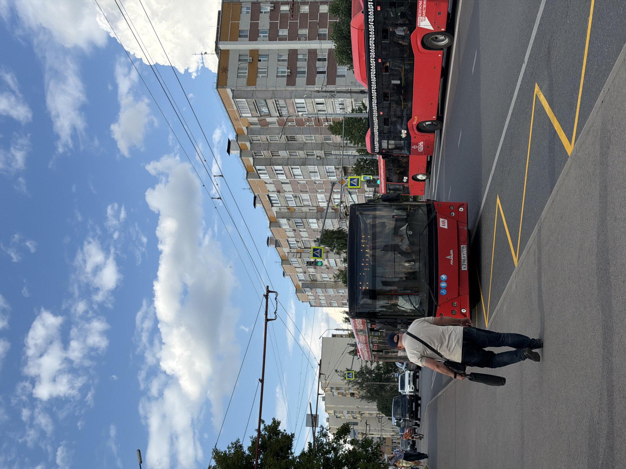 The media file shows a street scene with several red buses. In the foreground, a person is walking on the sidewalk, carrying a bag and wearing a light-colored t-shirt, dark jeans, and a baseball cap. Behind the person, a black bus is visible, and further back, several red buses are parked or moving. One of the red buses has text on its side, reading "x396y7116". The scene appears to be a city street with apartment buildings in the background. The sky is blue with scattered white clouds, suggesting it is daytime. The weather appears to be pleasant. Yellow lines are painted on the road, indicating a bus stop or a designated lane. The perspective of the image is from a slightly elevated angle, looking down at the street. The image is rotated 90 degrees clockwise.