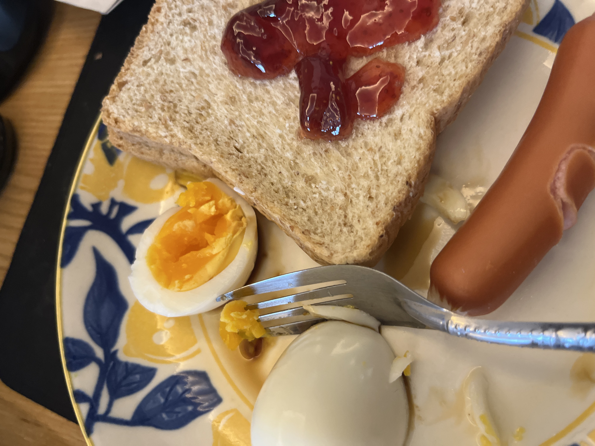 The image displays a close-up view of a meal, likely breakfast or brunch, served on a ceramic plate with a distinctive blue and yellow floral or foliage pattern. The plate rests on a light wooden table, with a dark placemat visible to the left.
The meal consists of several items. A slice of whole grain bread, topped with a generous dollop of shiny red strawberry jam, occupies the upper central part of the frame. To the right, a light brown hot dog or frankfurter is visible. Towards the lower left, a hard-boiled egg is cut in half, exposing its vibrant orange-yellow yolk. Another hard-boiled egg, mostly whole but partially cut, is situated below.
A silver fork is actively engaged with the food, its prongs inserted into the yolk of the halved egg, suggesting the meal is currently being consumed. A silver knife is also visible near the hot dog and the other egg. Small fragments of egg yolk are scattered on the plate.
The lighting indicates it is daytime. No people are visible, and there is no discernible text within the image. Based on the provided context, this meal is taking place in Pattaya City, Thailand.