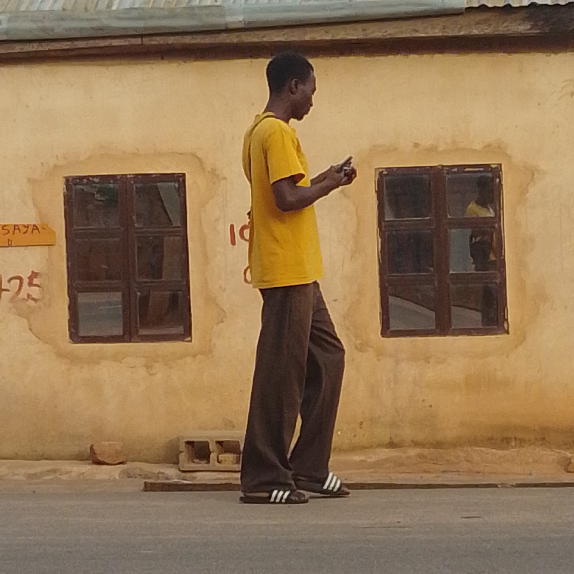 A dark-skinned man wearing a yellow t-shirt and dark brown pants is standing on a street in front of a building with a textured yellow wall. He is holding a mobile phone in his hands and appears to be looking at it. The building has two windows with dark frames and multi-pane glass. Red graffiti or writing is visible on the wall to the left of the man. The ground in front of the building is a paved road. The overall scene suggests an outdoor urban environment in Tempure, Nigeria.