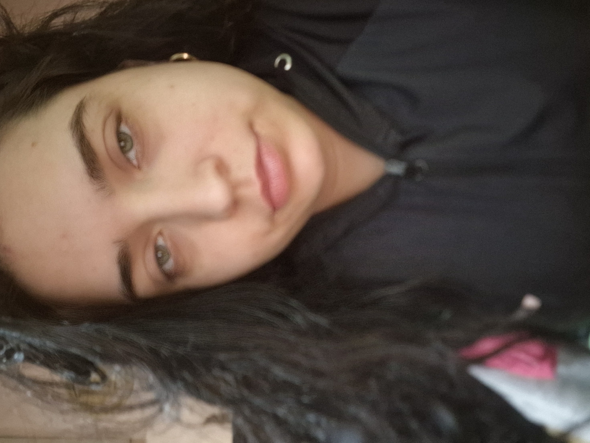 A young woman with dark hair and fair skin is lying down, her head tilted to the side. She is wearing a black hooded sweatshirt. Her eyes are open and looking at the camera. Her lips are slightly parted in a smile. A gold hoop earring is visible on her left ear. The background is out of focus.