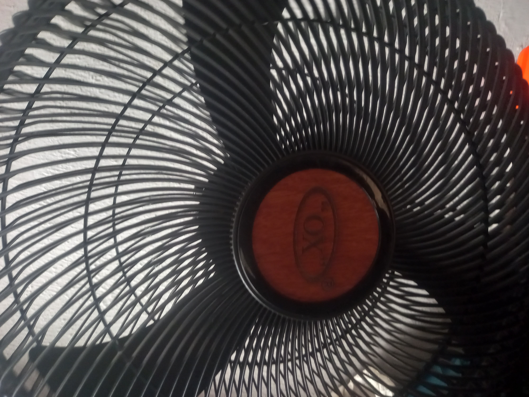 The image is a close-up, angled shot of a black oscillating fan. The fan has a circular grille with numerous thin, black bars arranged radially. The grille casts shadows on a textured white surface behind it, which appears to be a wall. In the center of the fan is a circular emblem with a reddish-brown wooden texture and a visible logo. The logo appears to be embossed and reads "FOX" in a stylized font, with a smaller circular symbol above and below the word. The fan is stationary, and no people are visible. The lighting suggests an indoor setting, possibly during the daytime due to the shadows. The overall impression is that of a functional household object. The provided location context is Ilado, Nigeria, which suggests the fan is located in this region.