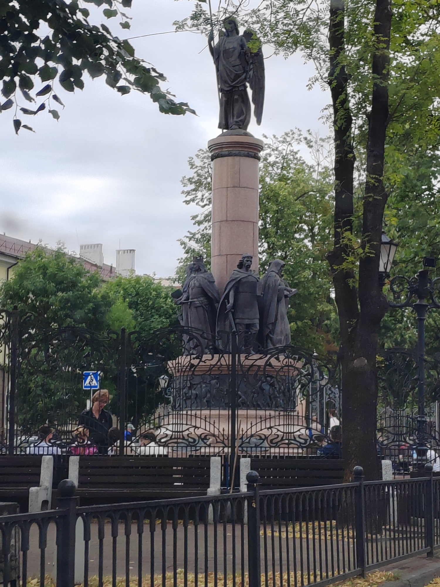 A monument with a statue of an angel on a tall column stands in a park in Brest, Belarus. At the base of the column are three statues of men in robes. In the foreground, there is a black metal fence, and behind it, a wooden bench with several people sitting on it. A woman in a black jacket is standing near the bench, looking at her phone. The trees are green and full of leaves, indicating it is likely summer or early autumn. The sky is overcast.
