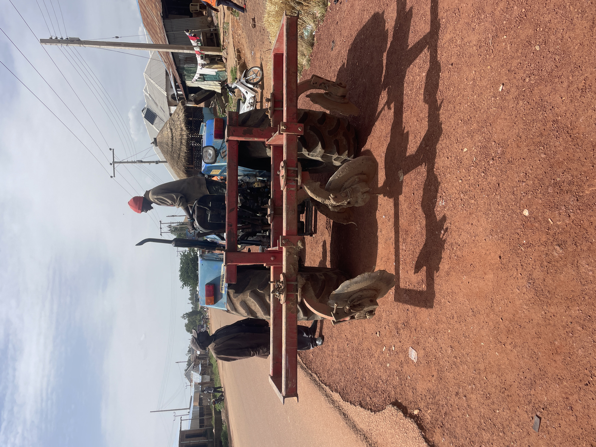 The image shows a blue New Holland tractor with a red plow attachment parked on a red dirt road in Durumi, Nigeria. Two men are standing near the tractor; one is wearing a red cap and the other a dark jacket. There are buildings, power lines, and a motorcycle visible in the background. The scene is bathed in sunlight, casting shadows of the tractor and plow on the ground.