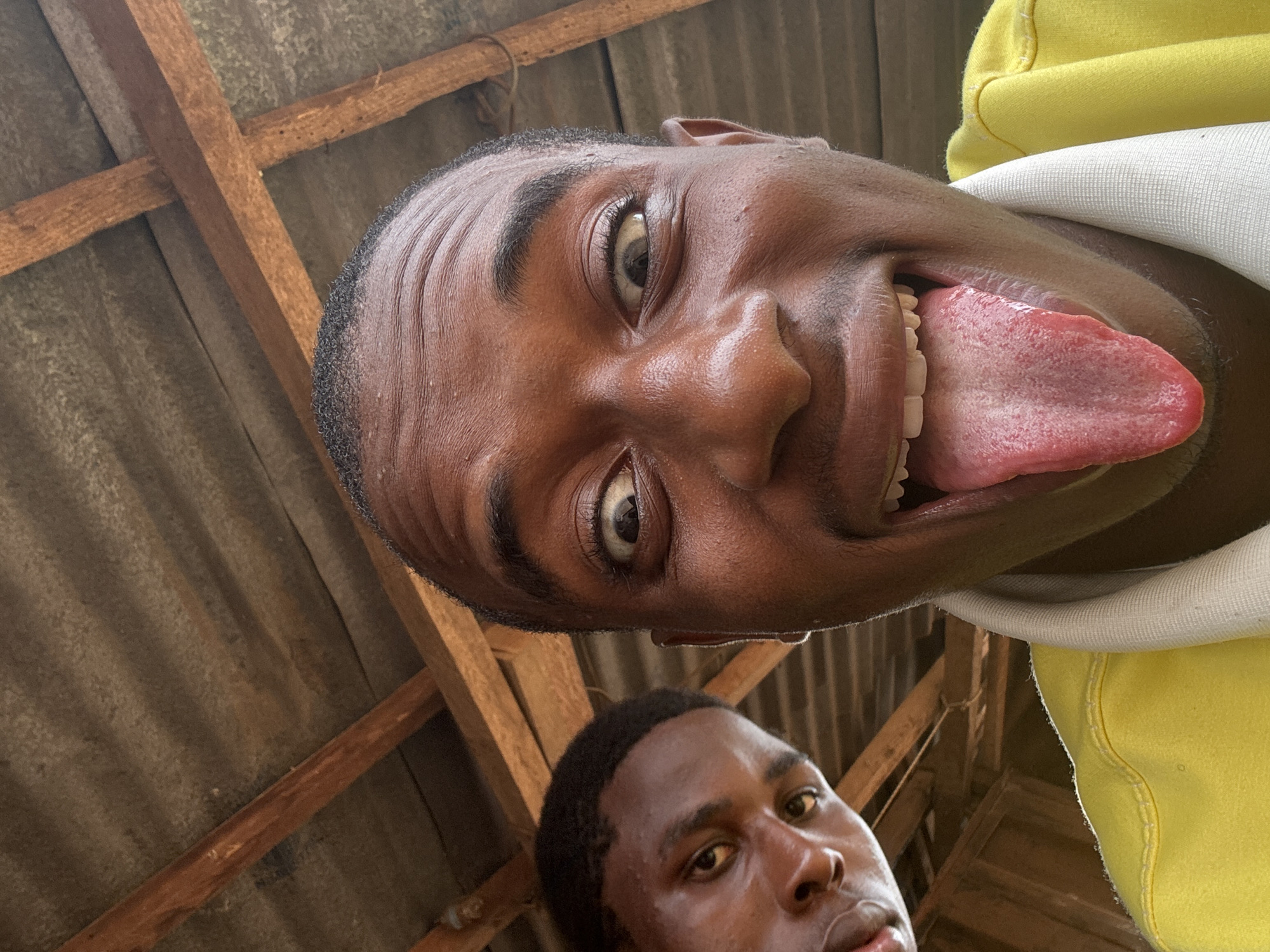 Two young Black men are in a selfie, looking up towards the camera from a low angle. They are under a wooden beam and corrugated metal roof structure. The man in the foreground is on the right, with his face close to the camera. He has his eyes wide open and is sticking out his tongue. He is wearing a bright yellow shirt with a white collar. The second man is on the left, further back and slightly out of focus. He is looking to his left with a more serious expression. The setting appears to be an outdoor or semi-outdoor space with visible wooden beams and a rough, textured ceiling. This scene is likely located in Nkpor, Nigeria, given the context.