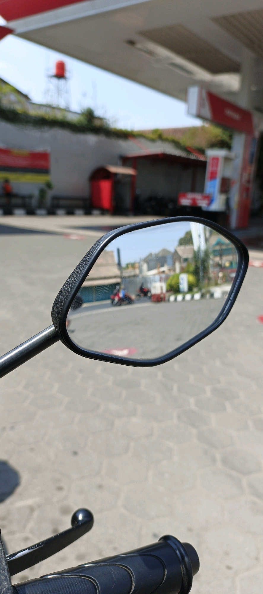 The image captures a reflective view in a motorcycle's side mirror, showing a street scene in Kasihan, Indonesia. The mirror reflects buildings, houses with tiled roofs, and two motorcycles, one ridden by a person wearing blue. The foreground is dominated by the motorcycle's black handlebar and the side mirror. Behind the mirror, a paved area with hexagonal tiles is visible, and further back, structures resembling a gas station canopy, a red booth, and a red water tank on a platform are present. The sky is clear and blue. The scene appears to be an outdoor public space on a sunny day.