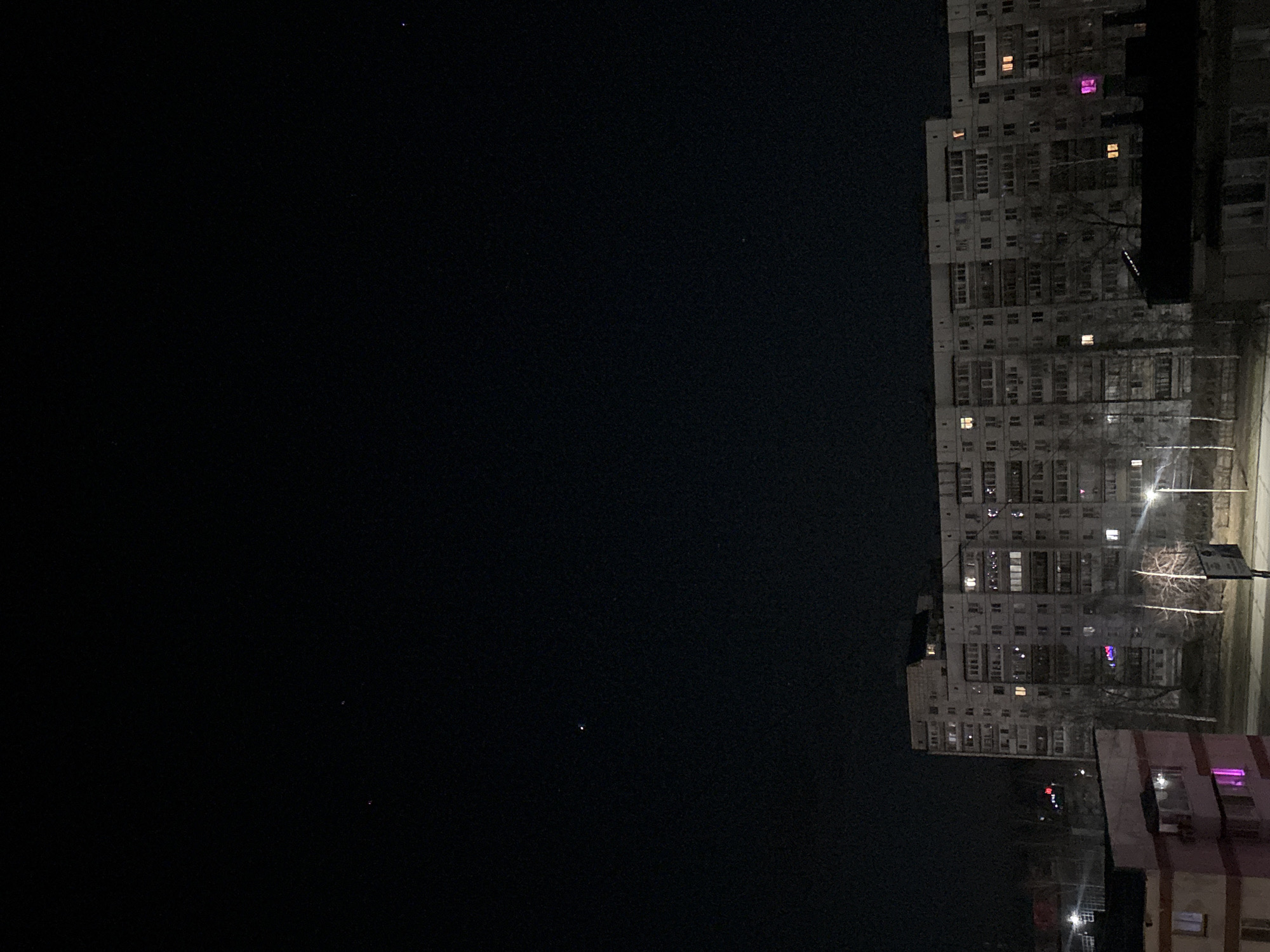The image shows a nighttime scene in what appears to be a residential area. There are several apartment buildings visible, some with lights on in the windows. The sky is dark, suggesting it is late at night. The location is likely Chaikovsky, Russia. There's a street light illuminating a small patch of land in front of one of the buildings. The overall atmosphere is quiet and still.