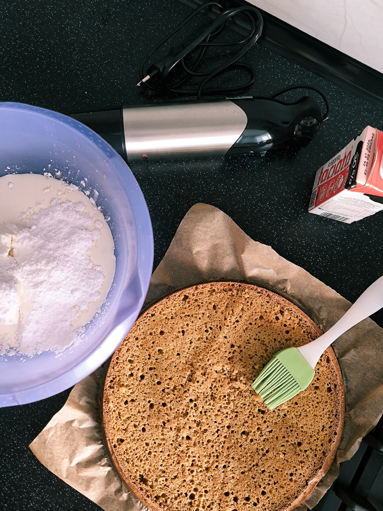 The scene depicts a kitchen countertop in Vinnytsia, Ukraine, showing various items associated with baking. A dark, speckled countertop serves as the primary surface. In the upper left, a light blue plastic bowl contains a white creamy liquid partially covered by a mound of white powdered substance. Centrally, a silver and black immersion blender with a coiled black power cord lies horizontally on the counter. To its right, a red and white cardboard carton, labeled "Łaciate" and "śmietanka" (Polish for cream) with an expiry date partially visible as "26.07.2026," rests on the counter. In the lower right, a round, light brown sponge cake layer with a porous texture is positioned on crumpled brown parchment paper. A white-handled silicone pastry brush with light green bristles rests on the surface of the cake layer. No individuals are present in the image.
