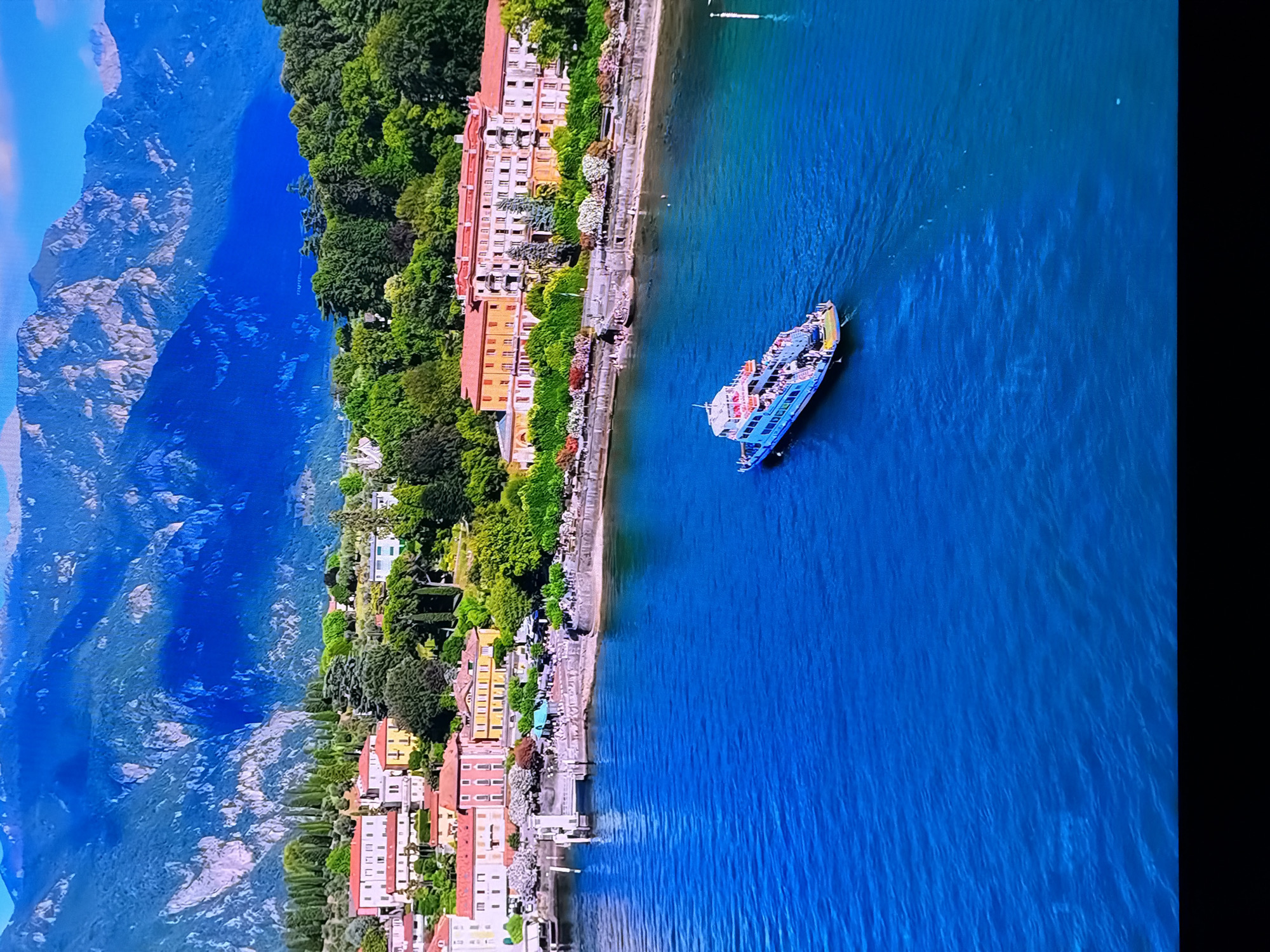 An aerial view captures a vibrant scene on a body of water, likely a lake, with a town nestled along its edge. Majestic mountains, shrouded in a deep blue hue, form a dramatic backdrop to the picturesque landscape. The town features a collection of buildings with red-tiled roofs, interspersed with lush green trees and flowering bushes. A prominent ferry boat, painted a light blue and white, cruises across the water, carrying passengers. The water itself is a striking, deep blue, reflecting the clear sky above. The image does not depict the city of Chirchik, Uzbekistan, or any recognizable landmarks from that location.