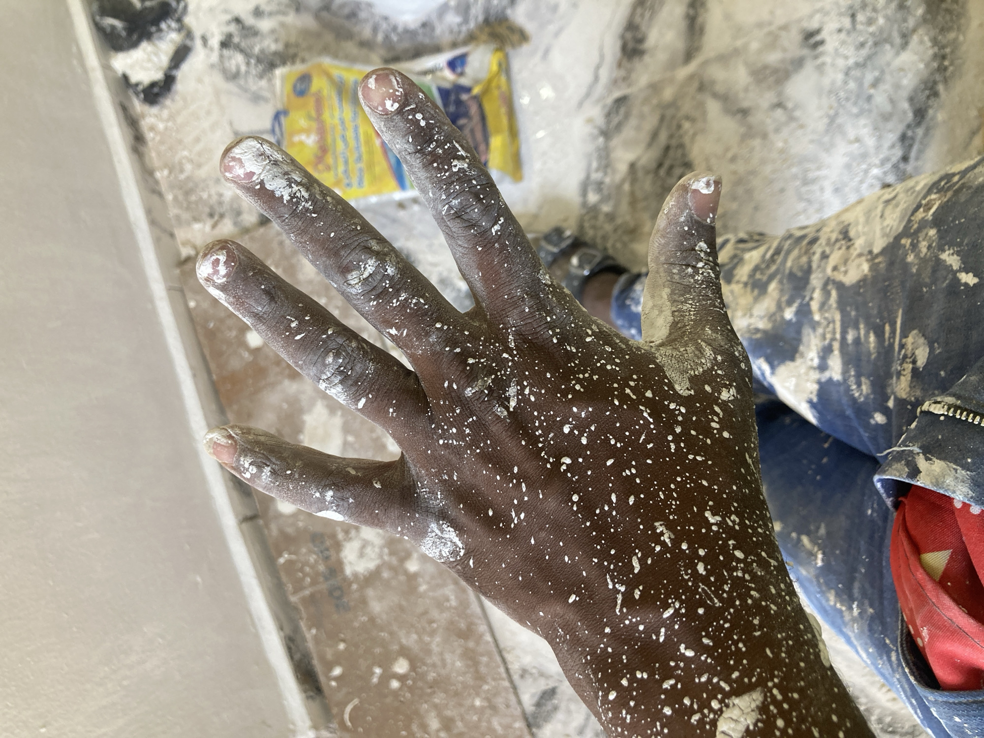 A close-up shot captures a person's left hand, which has dark-toned skin, heavily splattered with dried white material. Numerous small white speckles cover the palm and all fingers, including the fingernails, suggesting involvement in an activity such as painting, plastering, or whitewashing.
In the background, part of the person's body or clothing is visible, featuring blue denim-like fabric, also extensively stained with the same white substance. A splash of red fabric, possibly a lining or accessory, can be seen peeking out from a pocket area. The surrounding environment appears to be a worksite or a messy space, with white dust or dried material covering surfaces. A yellow packet or bag with faint, unreadable dark text is situated in the upper background amidst the scattered debris. No other text is clearly discernible anywhere in the image due to blurriness and visual obstructions. The image provides no specific cues about the time of day or external weather conditions, implying an indoor or covered setting for the activity.