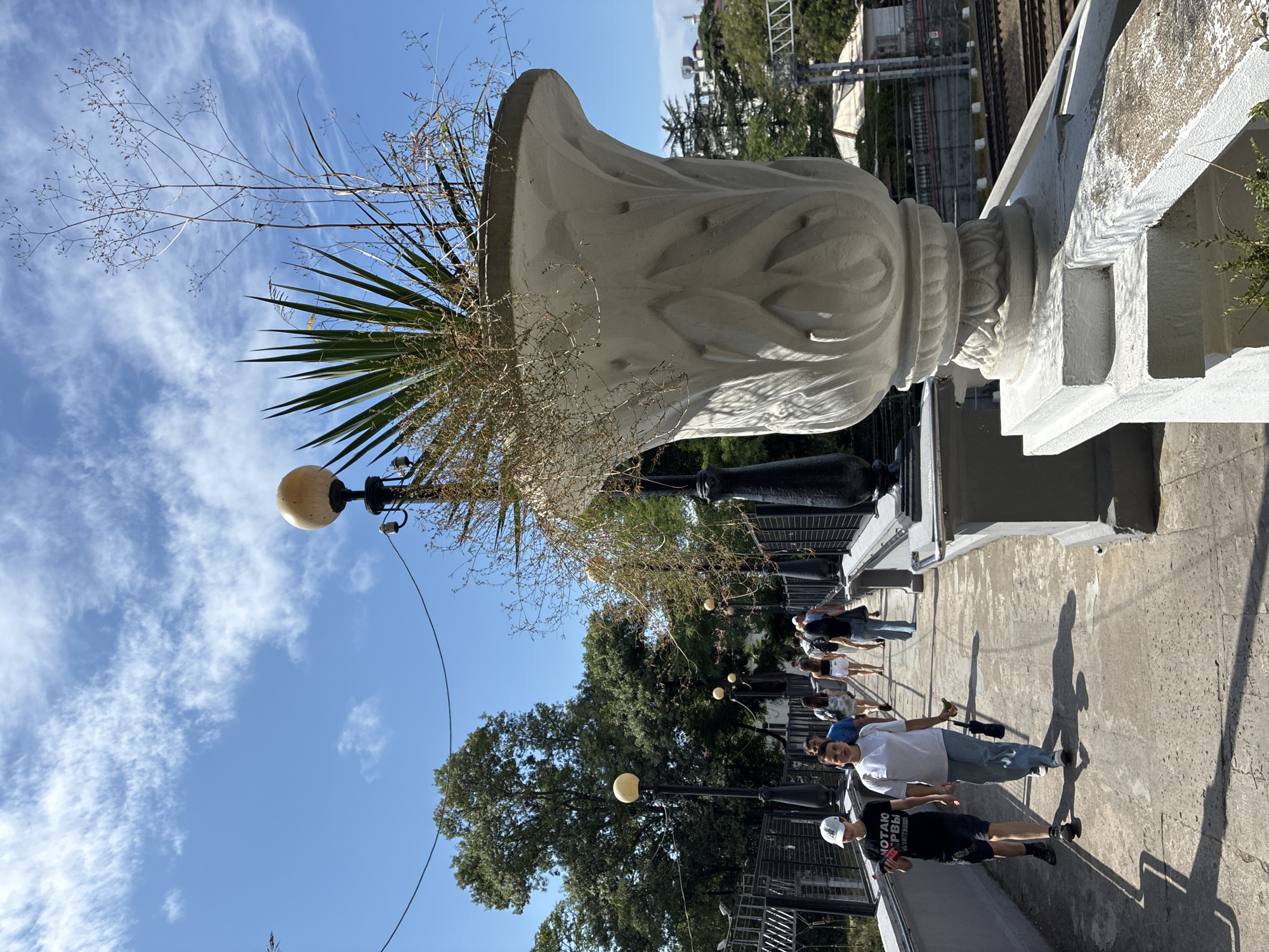 The image captures a sunny day on a pedestrian bridge in Sochi, Russia. A group of people are walking across the bridge, some heading towards the viewer and others away. The bridge is lined with ornate lampposts and large decorative planters. One prominent planter, in the foreground, holds a spiky green plant and dry, wispy foliage. Beyond the bridge, trees and buildings are visible, with train tracks running alongside the area. The sky is blue with scattered white clouds.
