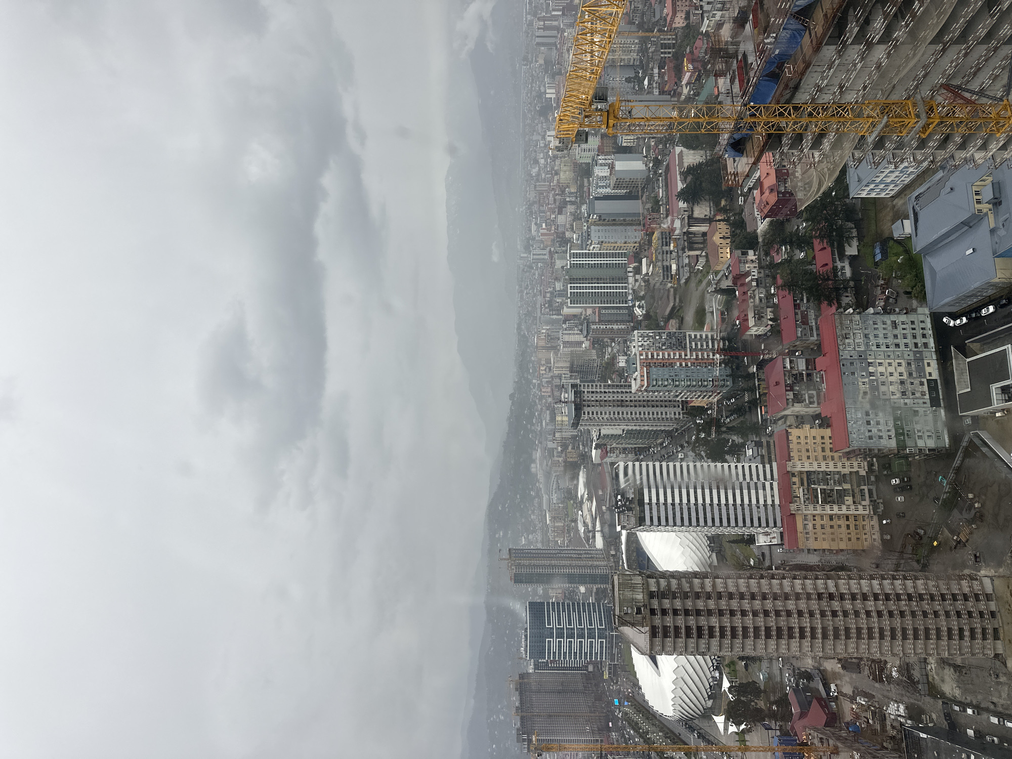 The image captures an aerial view of the city of Batumi, Georgia, taken on a cloudy day. A portion of the sky filled with heavy clouds occupies the left side of the frame. The cityscape is densely packed with buildings of varying heights and architectural styles. Visible construction cranes indicate ongoing development projects. Towards the lower part of the frame, there are several large buildings under construction. Numerous cars and some scattered trees are visible.