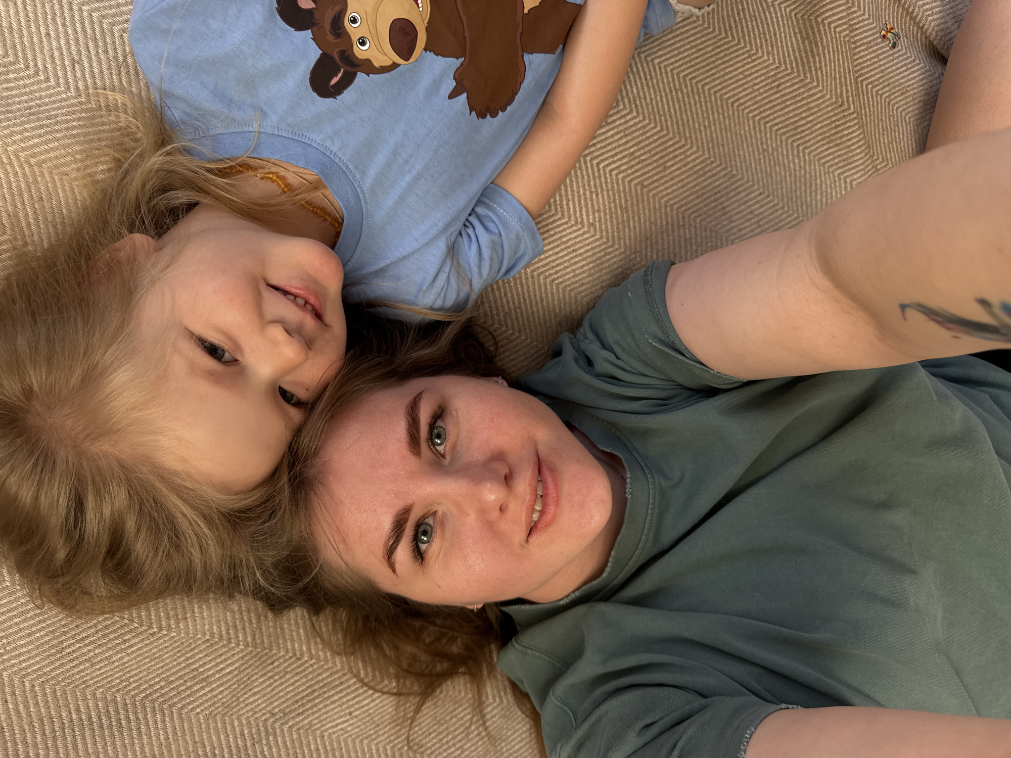 The image shows two individuals lying on a patterned beige surface. The individual on the left is a young child with blonde hair, wearing a light blue t-shirt with a cartoon bear on it. The individual on the right is an adult with light brown hair, wearing a darker green t-shirt, with what appears to be a tattoo on their arm. Both are looking towards the camera with smiles. The surface they are lying on appears to be a patterned rug or carpet. The location is Ферма, Russia, according to user input, though no elements in the image can verify this detail.