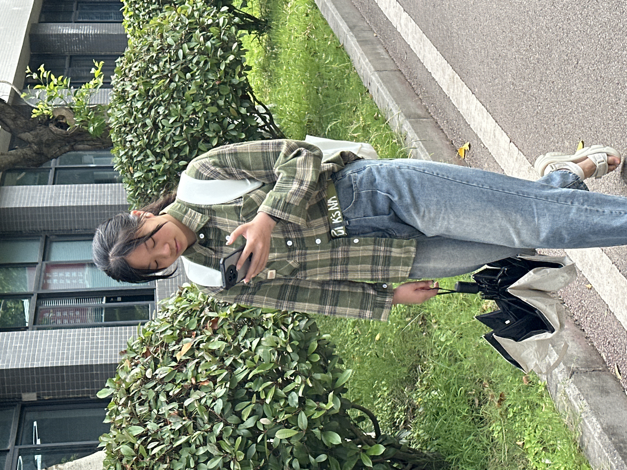 A young Asian woman stands in an outdoor scene in what appears to be Jiangjin, China. She is holding a smartphone and looking down at it. She has dark hair pulled back, and is wearing a green and tan plaid shirt, blue jeans, and white sandals. She has a white backpack on her back and an olive green belt with the word "UNK NA" on it. She holds a closed umbrella in her left hand. She is standing on a sidewalk next to a street with white lines. Adjacent to the sidewalk is a grassy area with green bushes. In the background, a multi-story building with windows and gray tiled exterior is visible. The scene is bathed in natural daylight.