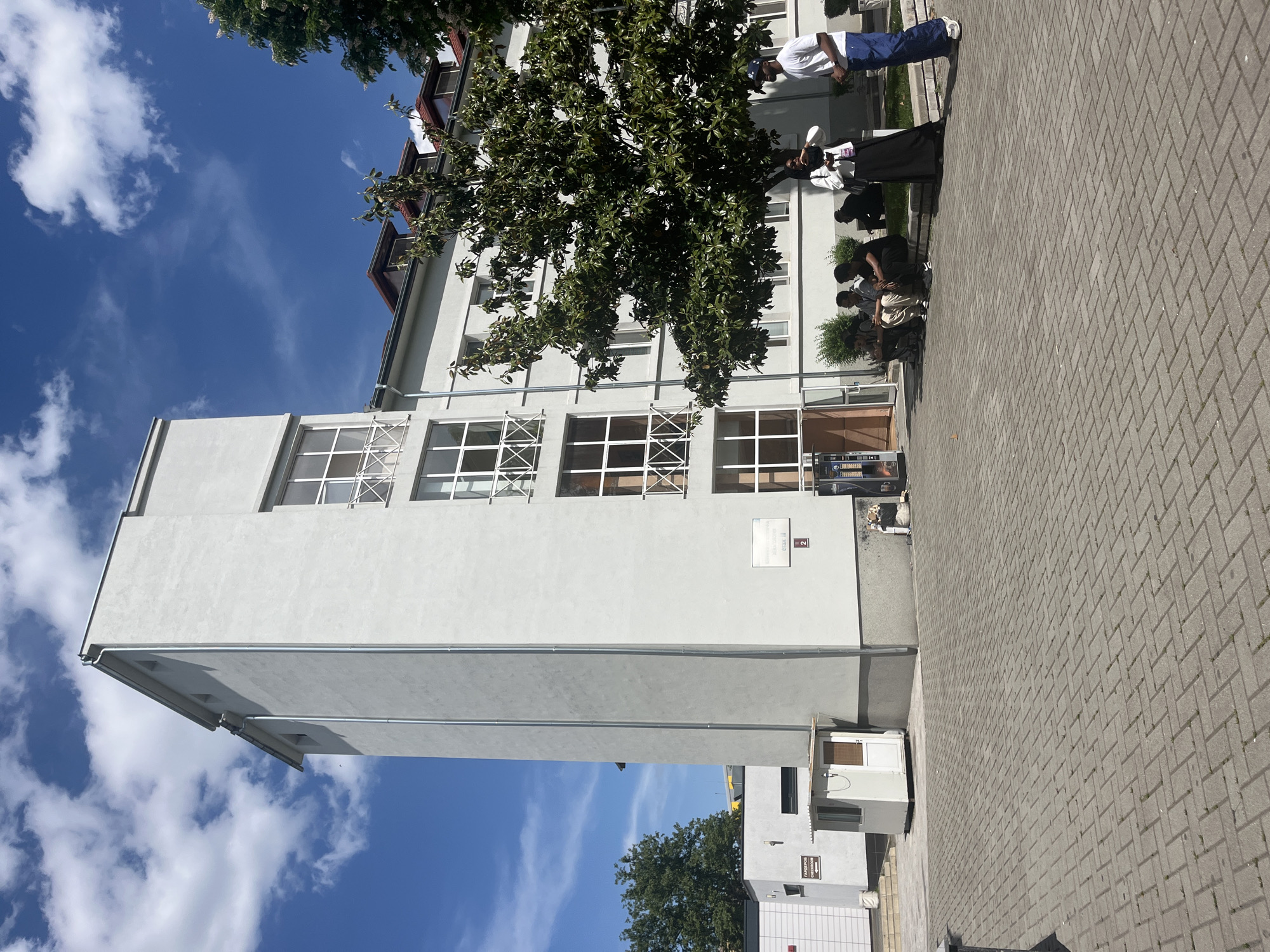 The image captures a view of a modern, boxy white building in Plovdiv, Bulgaria, under a bright blue sky with scattered clouds. A large green tree partially obscures the building's facade, which features several large, rectangular windows with white frames and cross-bracing. A group of people, mostly dressed in dark clothing, are gathered on a paved area adjacent to the building, some seated and some standing. The pavement is composed of grey rectangular blocks. A small white structure is located in the foreground to the right of the frame.