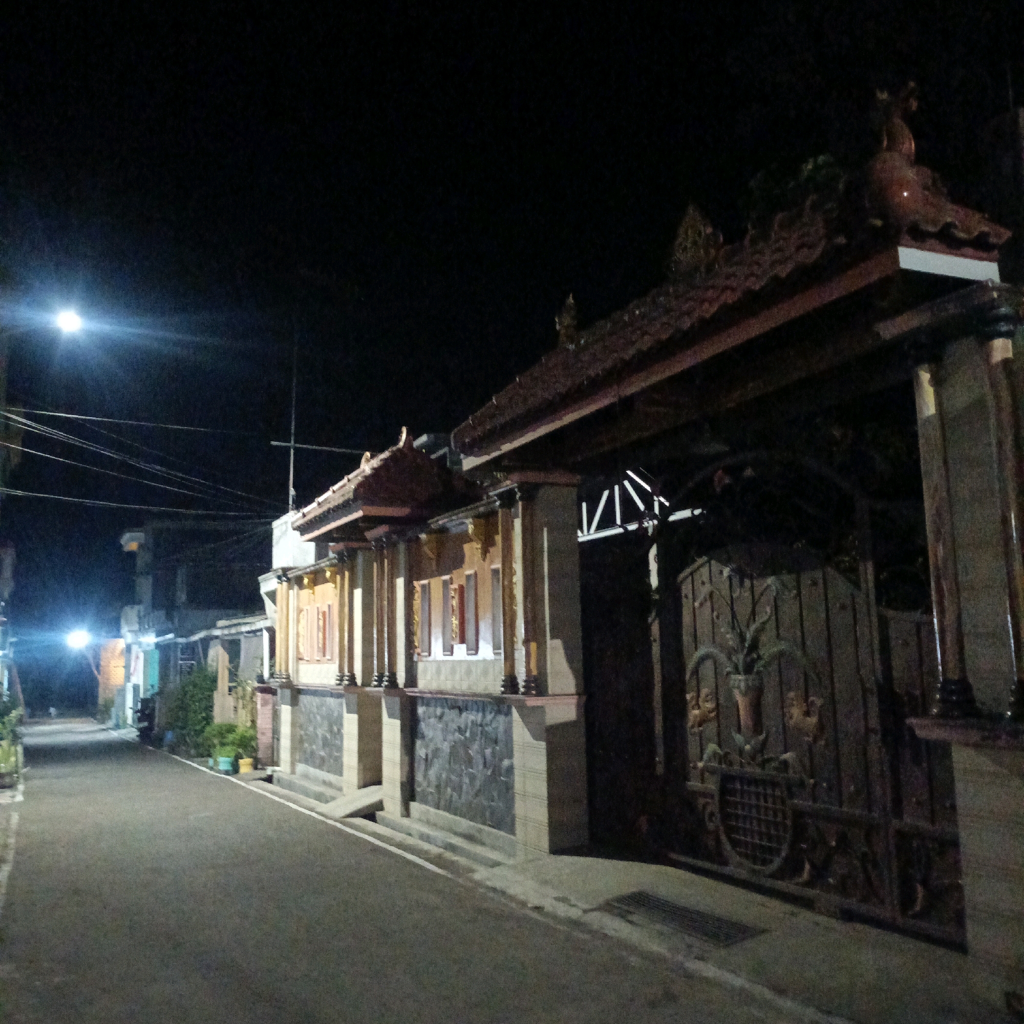 An outdoor, low-light, eye-level, medium shot captures a street scene at night in Salatiga, Indonesia. The street is paved and curves to the left, illuminated by a streetlamp on the left side of the frame and lights from buildings. A building with traditional Balinese-style architecture stands on the right side of the street, featuring a decorative roof, columns, and a large, ornate metal gate. The gate has intricate patterns, including a plant motif in a pot. The building's facade is textured with stone and has several windows with reddish frames. Power lines crisscross the dark sky above. The overall impression is one of a quiet residential street at night, with a prominent, artistically designed entrance.