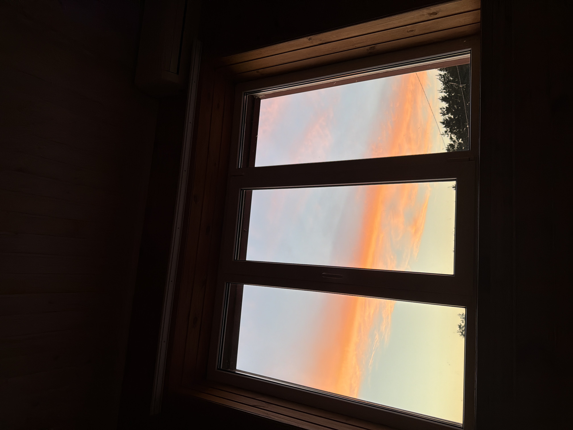 This is a photograph of a three-pane window looking out at a sunset. The sky is a vibrant mix of orange, pink, and pale blue, with wispy clouds. Below the sky, a dark silhouette of evergreen trees lines the horizon. The window frame is white, and the interior walls around the window are paneled with dark wood, suggesting the photo was taken from inside a room with a sloped ceiling, possibly an attic or a room in a wooden house. The scene is dimly lit from within the room, highlighting the bright sky outside. The photograph does not contain any people, animals, or specific identifiable landmarks that would indicate a location in Nizhnekamsk, Russia.