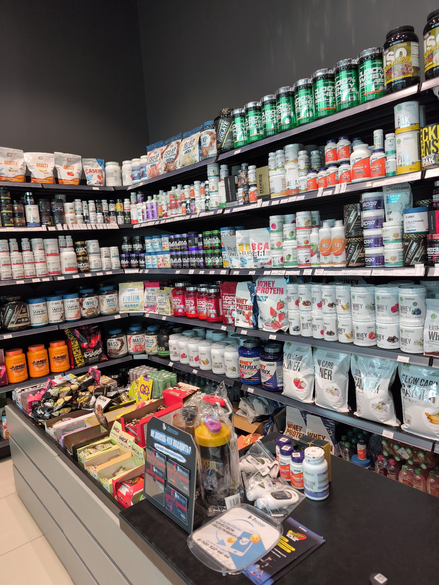 The image shows the inside of a nutrition supplement store. The store is brightly lit, with shelves stocked with various protein powders, vitamins, and supplements. The shelves are lined with containers of different shapes, colors, and sizes. In the foreground, there is a checkout counter displaying items such as snack bars, gift cards, a shaker bottle, supplements, and electronic devices. The wall behind the shelves is a dark gray. The scene appears to be in Moscow, Russia, based on the text on the gift card display, which reads "Не знаешь что подарить?" (Don't know what to gift?).