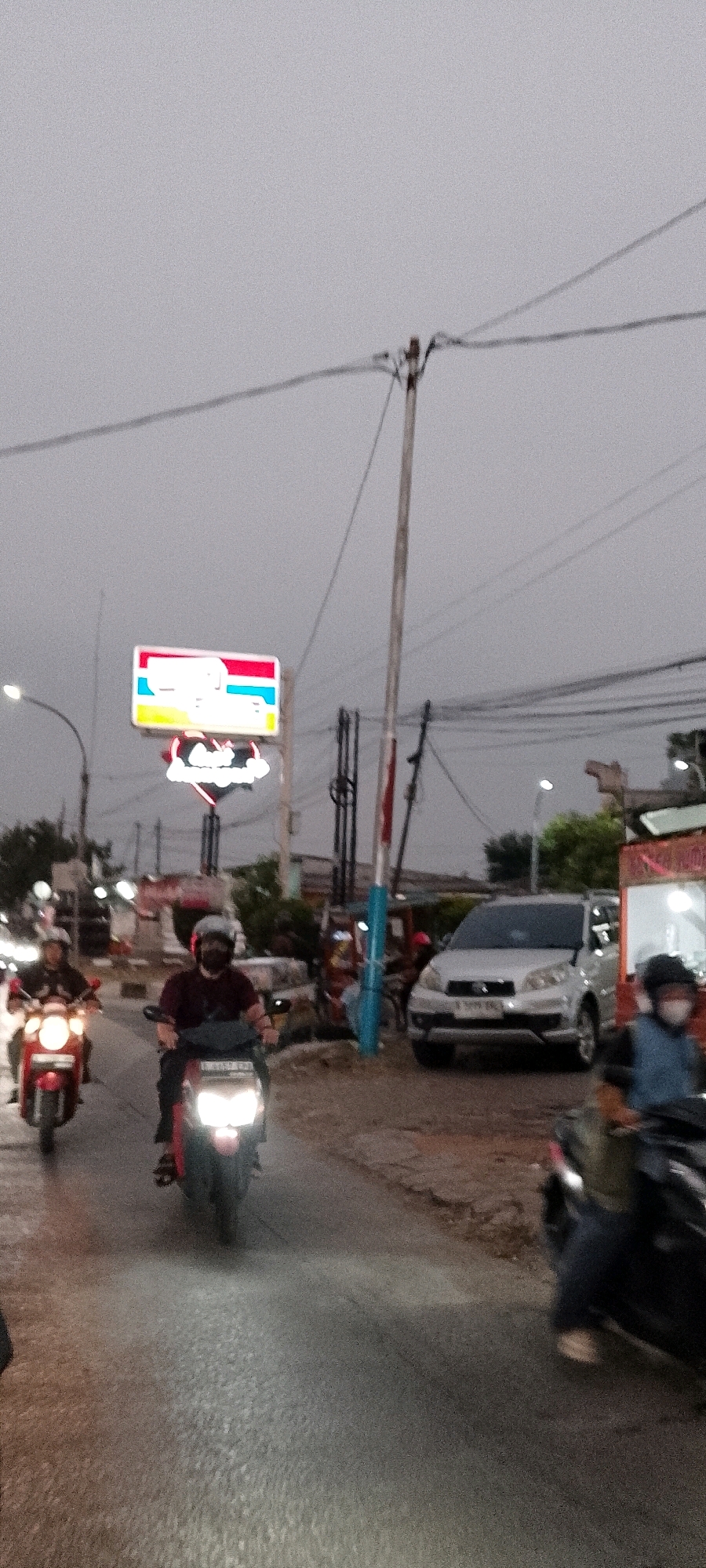 The image depicts a street scene in Depok, Indonesia, under an overcast sky. In the foreground, several motorcycles are visible on a paved road. One motorcycle in the center is illuminated by its headlights, with a rider wearing a dark shirt and helmet. To the left, another motorcycle with a rider in a dark jacket is also lit by its headlights. Further back on the right, a silver SUV is parked or stopped. A prominent sign with a red, white, blue, and yellow design is displayed on a pole, and below it, a heart-shaped neon sign with red lettering is illuminated. Overhead, electrical wires crisscross the sky, supported by utility poles. The environment suggests a commercial or roadside area with some buildings and foliage present in the background. The lighting indicates it is either dawn or dusk, or a heavily clouded day.