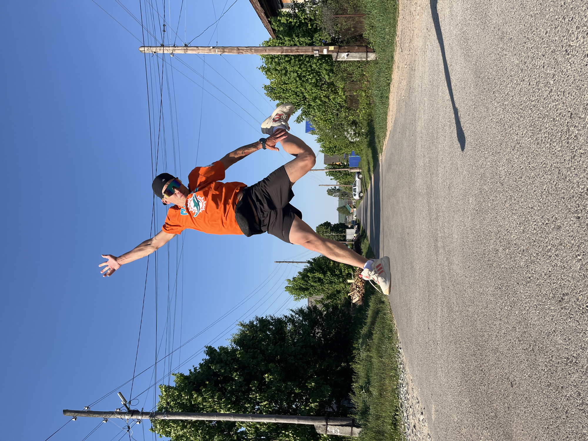 A person wearing an orange Miami Dolphins shirt, black shorts, a black baseball cap, and sunglasses, is pictured on a street in Chaikovsky, Russia. The individual is mid-air, with one leg extended and the other bent upwards. The person's arms are also extended. The street is paved and bordered by grass and trees. Utility poles with wires are visible on both sides of the street. A building and parked cars can be seen in the distance. The sky is clear and blue.