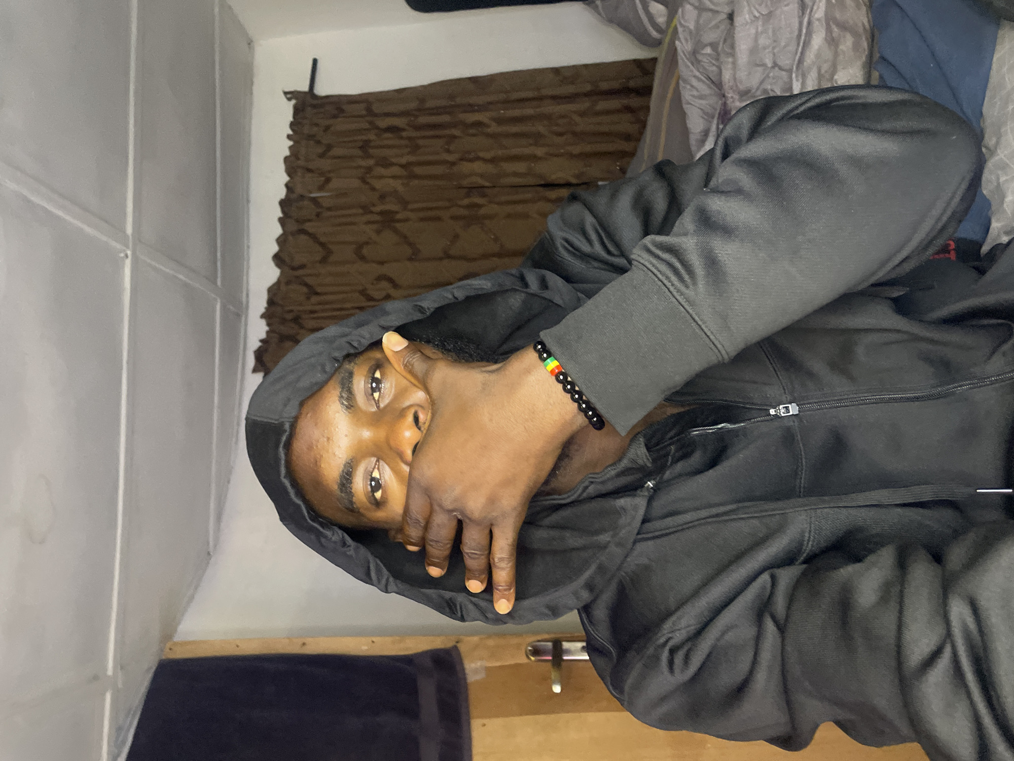 A man wearing a black hoodie covers part of his face with his hand. He is wearing a Rastafarian-colored bracelet. In the background are a white-paneled wall, a brown curtain, and bedding. The location is likely a private residence. There is no verifiable information to indicate that the image was taken in Jos, Nigeria.