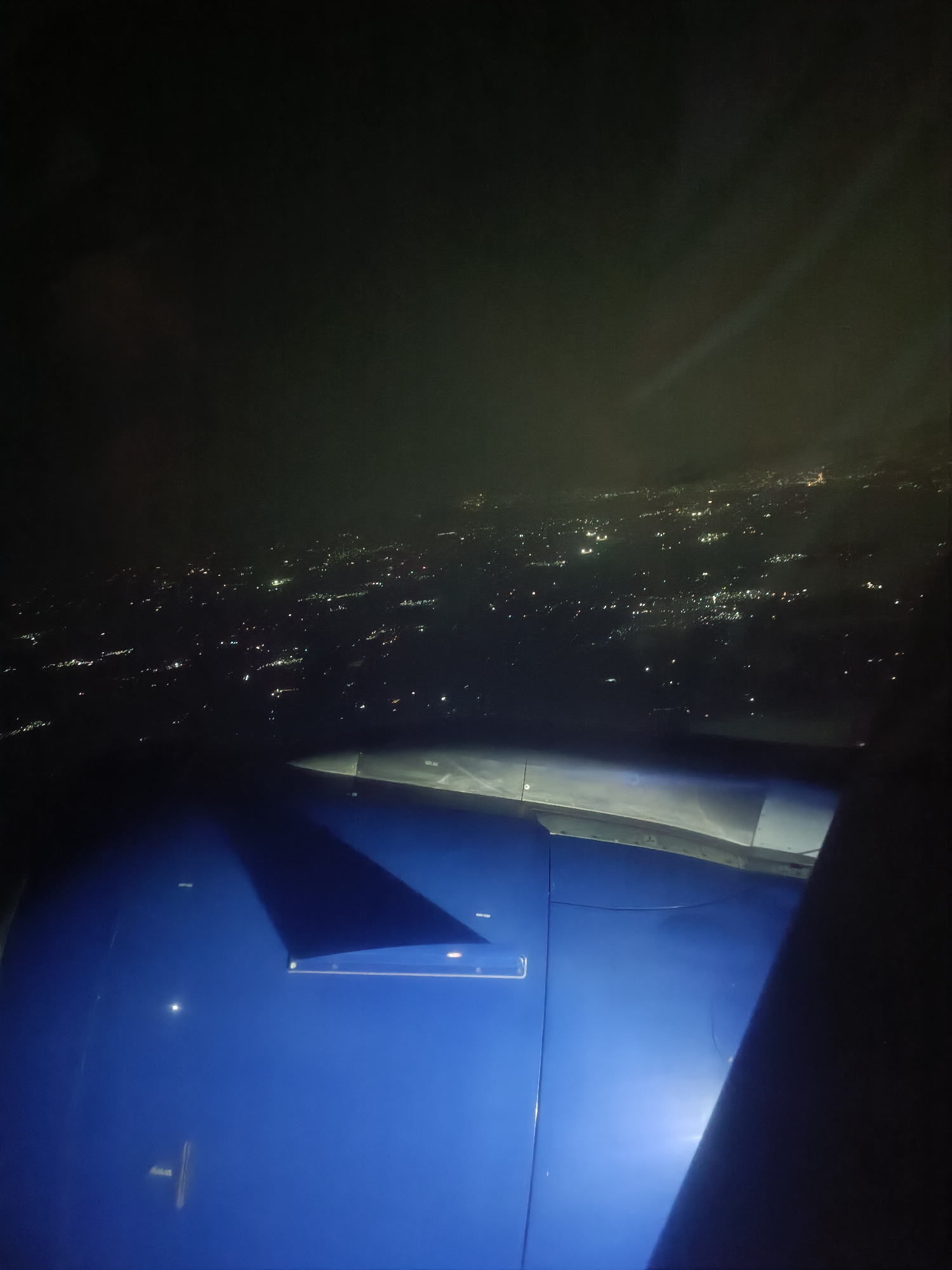 This is an aerial view of the city of Agassaim, India at night, as seen from an airplane window. The scene is dominated by the dark sky, with faint lights of the city twinkling below. Part of the airplane's wing and fuselage is visible in the foreground, painted a vibrant blue. The wing casts a shadow on the fuselage. The city lights appear as a dense pattern of small, white and yellow dots scattered across the dark landscape. Some areas of the city appear brighter, suggesting clusters of buildings or activity. The image captures the vastness of the urban sprawl seen from a high altitude.