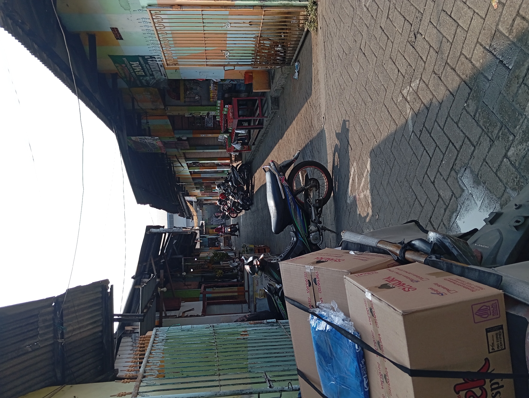 The image depicts a street scene in the city of Kepulungan, Indonesia, likely a market area, as suggested by the presence of stalls and vendors. The time of day appears to be morning or late afternoon due to the long shadows cast by the buildings and objects. The street is paved with interlocking brick pavers. On the left side of the street, there are buildings with corrugated metal roofs and a green metal gate. Several motorcycles are parked along the right side of the street and in front of the buildings. Some boxes are stacked on what appears to be a parked motorcycle in the foreground, one of which has text in a language other than English. The buildings on the right side of the street are painted in various colors, with some displaying banners and signs. One banner visible has the text "BMM". People are seen on motorcycles, and some are standing near the stalls. The overall atmosphere is that of a busy commercial street in a somewhat rustic setting.
